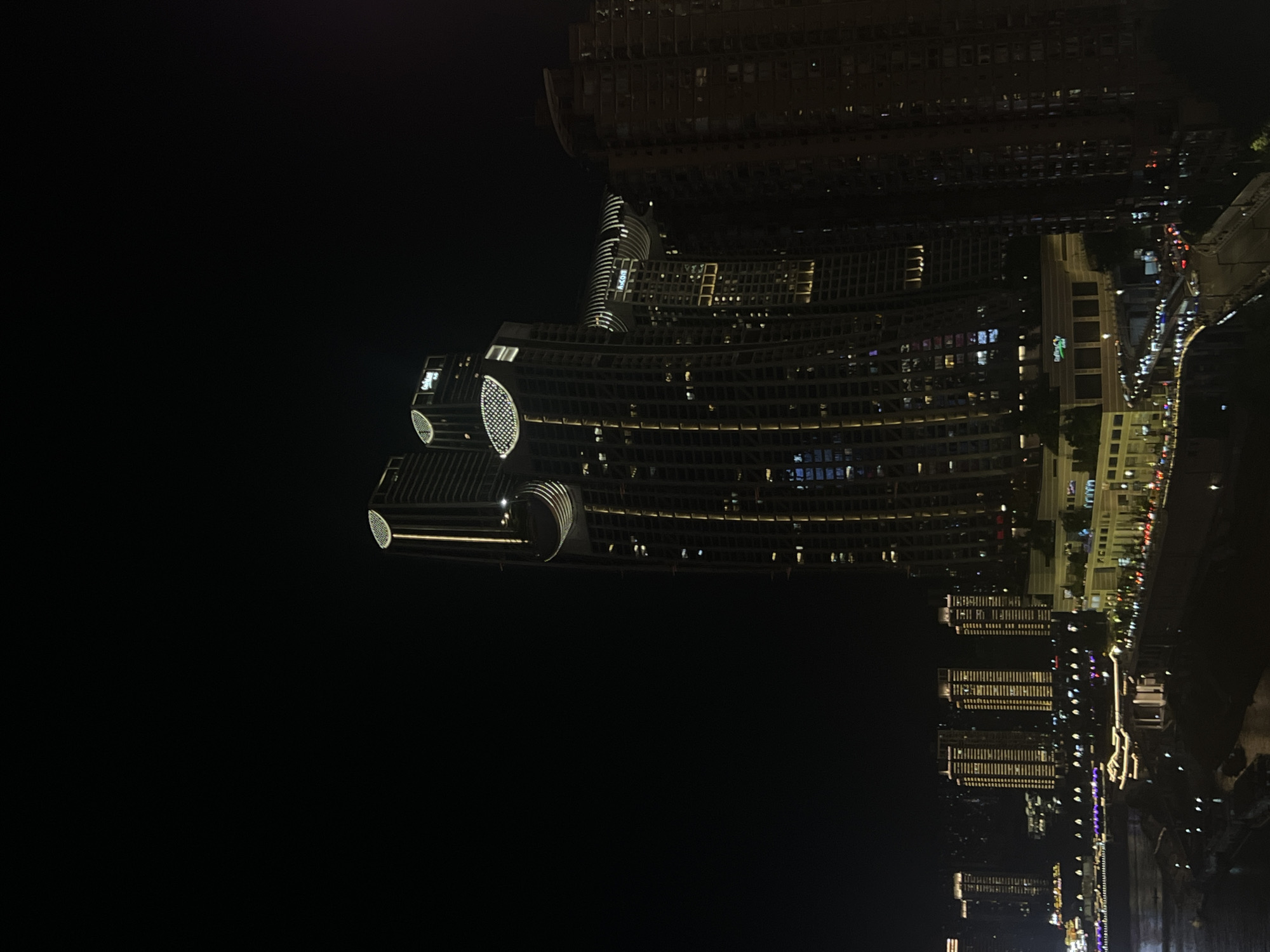 The image depicts a nighttime cityscape in Yuzhong District, China, characterized by illuminated skyscrapers and a waterfront. The main focus is on a cluster of modern high-rise buildings with distinctive architectural features, such as curved facades and illuminated upper sections. The buildings are lit up with a warm, yellow glow, highlighting their verticality and architectural details.
In the foreground, a river or body of water is visible, reflecting some of the city lights. Along the waterfront, there are lower-rise buildings and structures, some of which appear to be commercial or entertainment venues, also illuminated at night. A road or promenade runs alongside the waterfront, with visible car lights suggesting traffic.
The sky is dark, indicating it is night, with the lights of the city creating a contrast against the blackness. The overall impression is of a vibrant, developed urban environment at night, with a significant architectural presence. There are no discernible people or specific actions visible in the image. The scene is static and focused on the architectural landscape.