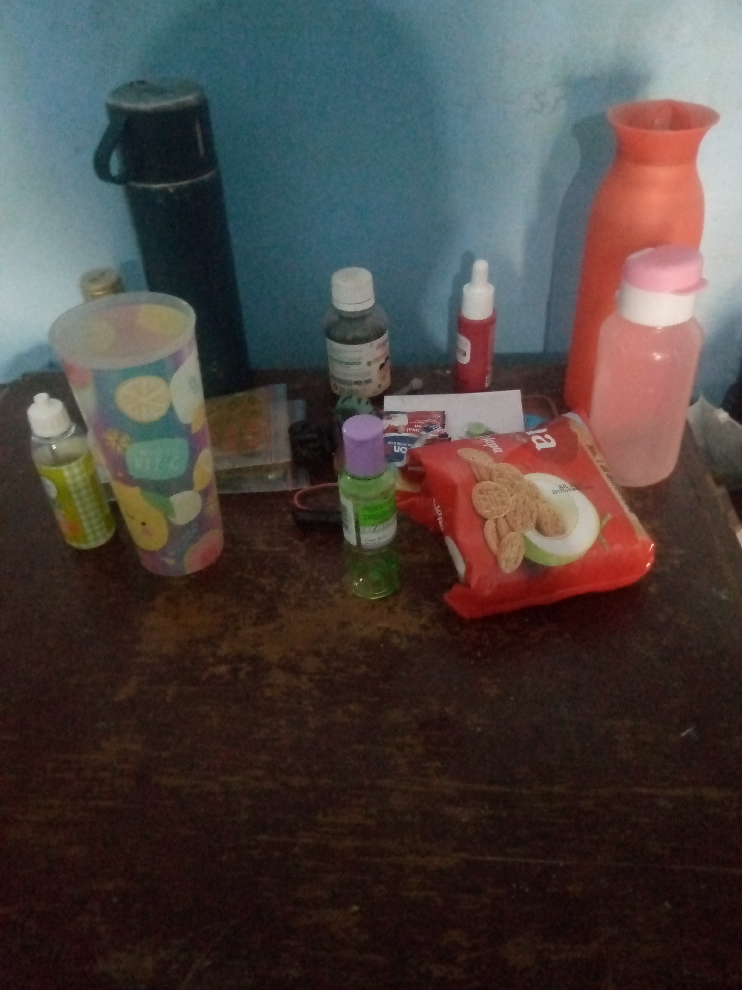 A collection of various bottles and a package of crackers are arranged on a dark wooden surface against a light blue wall. On the left is a tall, black thermos with a handle. To its right, a cylindrical container with a colorful fruit pattern is positioned. Next to that, a small, clear bottle with a yellow checkered label and a white cap is visible.
In the center of the arrangement, a bottle with a white cap and a label that appears to be for medicine or supplements stands upright. To its right, a small red bottle with a white cap is present. Further to the right, a taller, clear plastic bottle with a pink lid contains a light pink liquid. Behind these, a large, orange vase with a wide opening is placed against the wall.
In the foreground, a red package of crackers with an image of sliced apples and circular crackers is prominently displayed. To the left of the cracker package, a small, green bottle with a purple cap is standing. The surface the items are on is a dark, textured wood, and the background is a plain light blue wall, possibly indicating an indoor setting such as a home or shop in Giriasih, Indonesia.