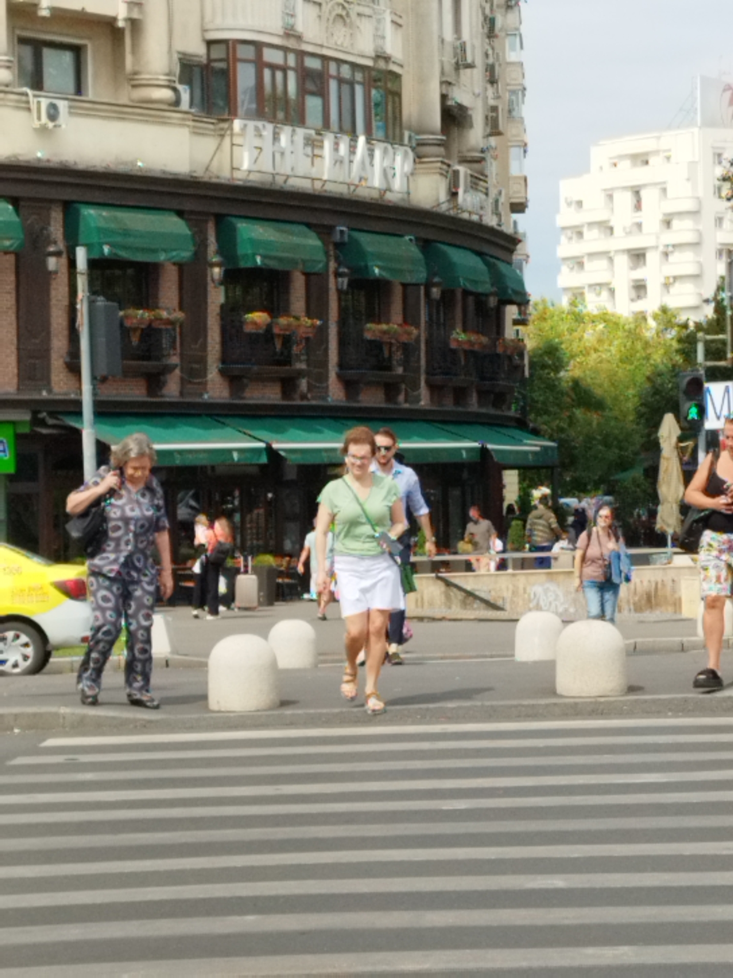 The image captures a street scene in Bucharest, Romania, near the Romanian Athenaeum. In the foreground, a pedestrian crossing is marked by grey and white stripes on asphalt. Several people are present on or near the crossing.
On the left, a woman wearing a patterned grey and black outfit with matching pants and a top, carrying a black backpack, is walking on the sidewalk. Beside her, a yellow taxi is parked.
In the middle ground, a woman in a green short-sleeved shirt and white shorts is crossing the street from right to left. Behind her, a man in a blue shirt and dark pants is walking towards the viewer. Further back, several other individuals are visible, some with luggage.
The background features a multi-story building with a dark facade and green awnings over its windows. Above the awnings, the building displays ornate architectural details and signage. Adjacent to this building is another white, modern-looking structure. Trees are visible behind the buildings. A traffic light showing green is present on the right side of the image. The overall scene depicts a bustling urban environment with everyday activity.