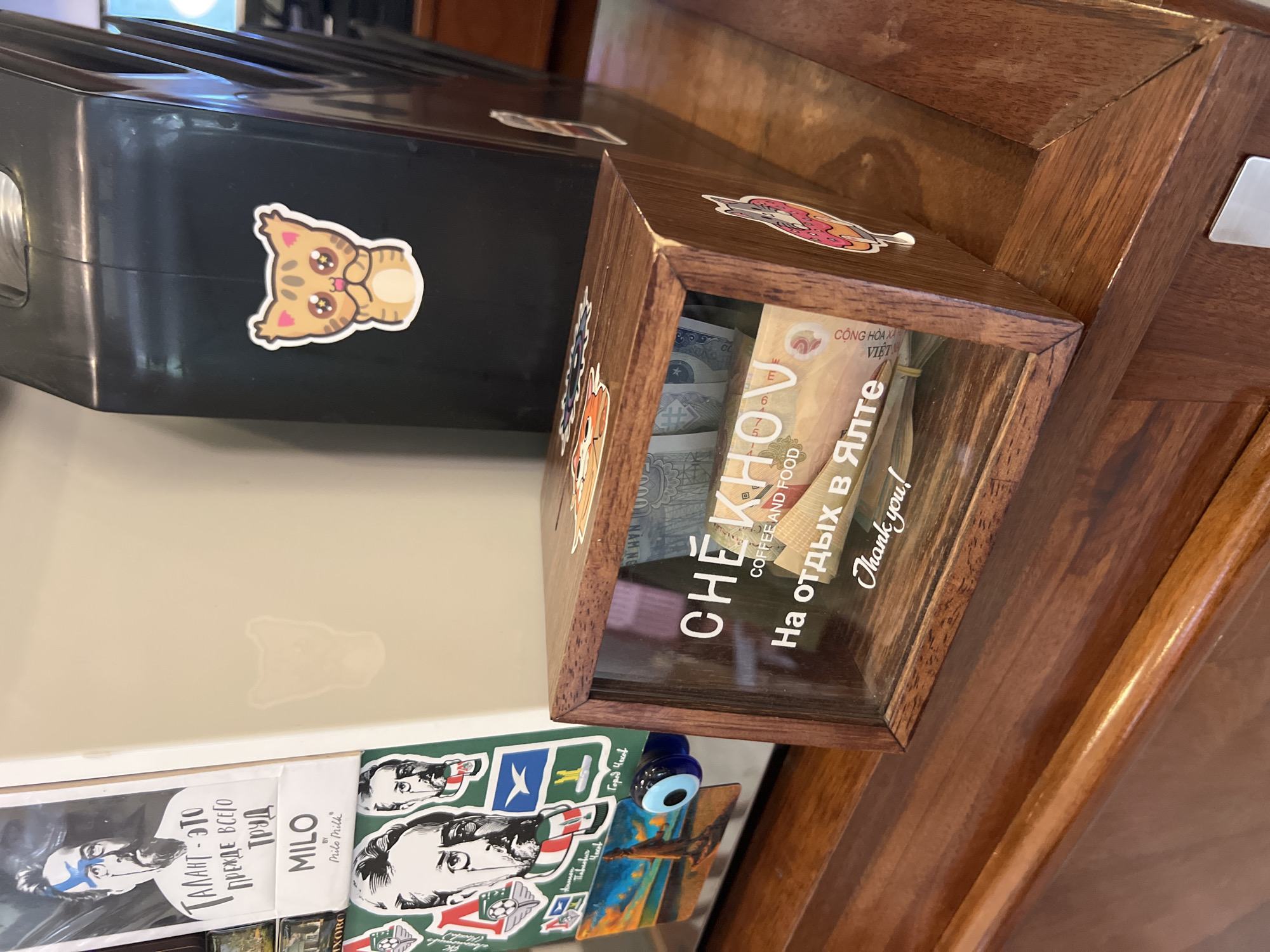 An indoor scene showcases a wooden tip or donation box with a clear front, filled with banknotes, sitting on a polished wooden surface. To its left is a black rectangular object, possibly an electronic appliance, adorned with a large sticker of a cartoon cat with sparkly eyes.
The wooden box, prominently labeled "CHEKHOV COFFEE AND FOOD," serves as a collection point. Written in Russian on its front are the words "На отдых в Ялте" (For a vacation in Yalta), indicating its purpose, followed by "Thank you!" in English. The banknotes inside appear to be Vietnamese Dong, with phrases like "CỘNG HÒA XÃ" and "VIỆT" visible on one bill. The box itself is decorated with various stickers.
Below these items, additional objects are visible, including what looks like books or pamphlets. One features a stylized portrait of a man with blue face paint and the Russian text "ТАЛАНТ - ЭТО ПРЕЖДЕ ВСЕГО ТРУД" (TALENT IS, ABOVE ALL, HARD WORK), along with "MILO BY Milo Milk." Other visible texts include "IXOBO" and "Город Чехов" (Chekhov City), further highlighting a Russian cultural presence. An "evil eye" bead and a small picture of a landscape with a flying object are also present. The overall setting suggests a casual, personalized atmosphere, likely within a cafe or shop in Nha Trang, Vietnam, a location known for its Russian community.