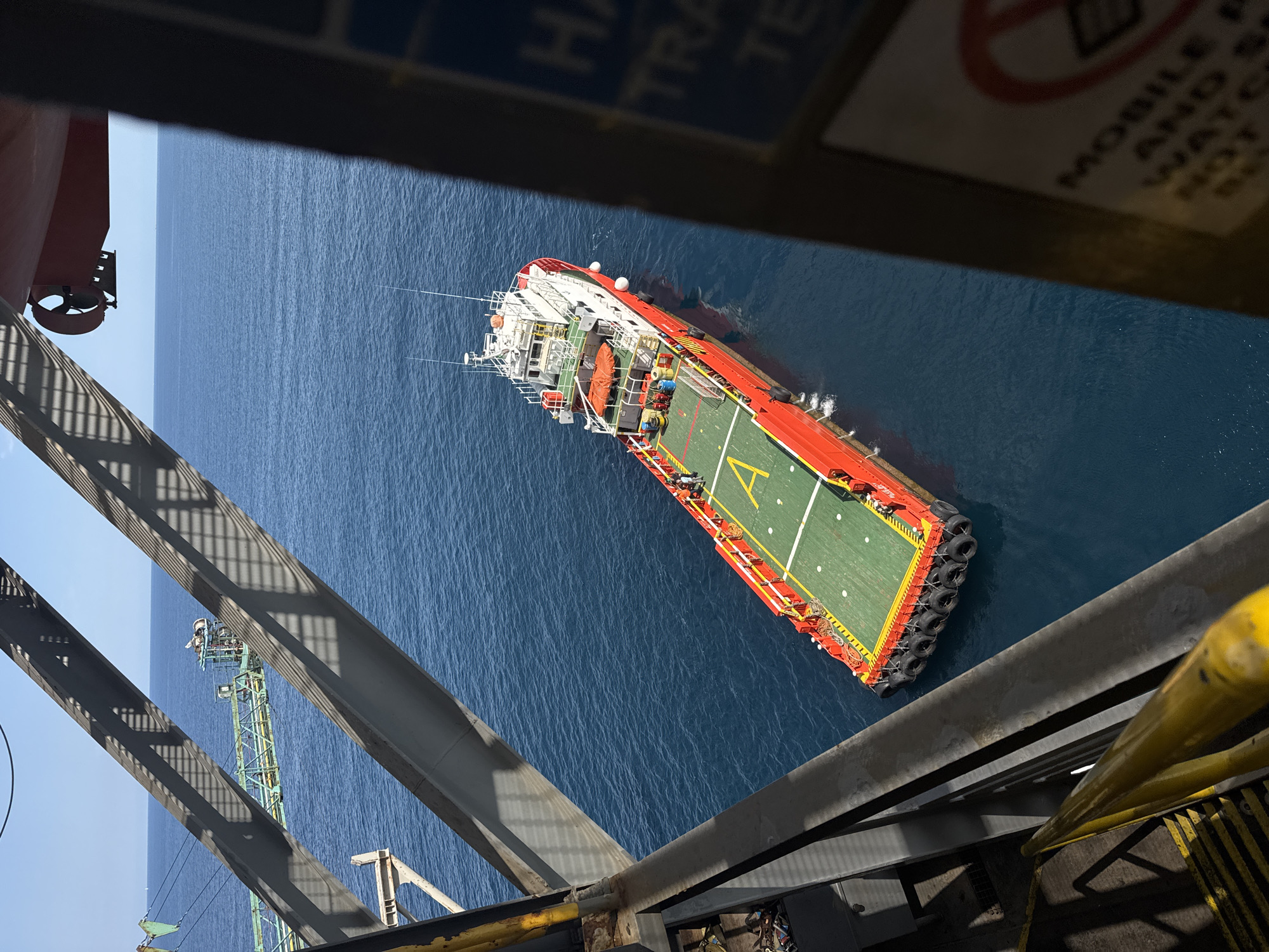 The image shows an aerial view of a red and white offshore supply vessel with a green helideck in a blue ocean. The vessel is positioned to the right of the frame, with its stern pointing towards the bottom right corner. The helideck features a large yellow 'H' marking. Several orange life rafts are visible on the vessel's deck, along with blue barrels and other equipment. A few people are visible on the deck of the vessel. In the background, the deep blue sea stretches to the horizon under a clear blue sky. Part of what appears to be a larger offshore structure or ship is visible in the foreground, framing the scene from the left and right. The foreground also shows metal beams and railings, suggesting the photo was taken from a raised platform. The scene is likely on an oil rig or a similar offshore platform in the open sea, not within a city.