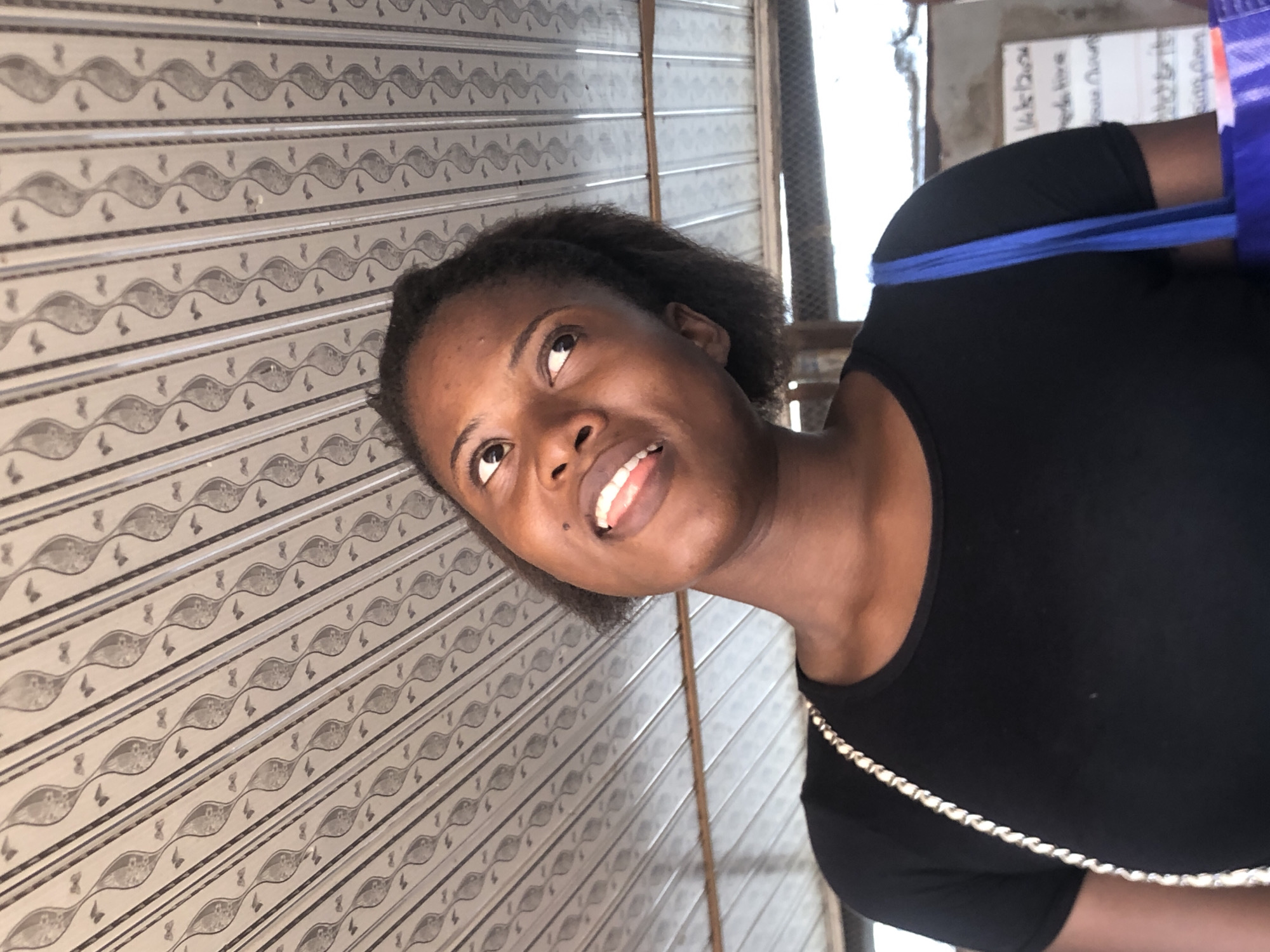 A woman with dark hair and a black shirt is standing in front of a textured wall. A blue strap is visible on her shoulder. The setting may be Ukwu-Abwa, Nigeria, though this is not directly confirmed by the image.