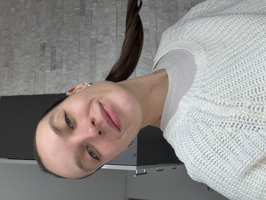 A close-up, chest-up shot captures a young woman looking directly at the viewer with a friendly, subtle smile. Her head is tilted slightly to her right. She has clear skin, bright blue-green eyes, and natural, well-defined eyebrows. Her lips are a soft pink.
Her long, dark brown hair is pulled back into a high ponytail, with the ponytail visible extending over her right shoulder (from the viewer's perspective). She is wearing silver-colored hoop earrings in both ears.
She is dressed in a cream or off-white knitted sweater featuring a distinct ribbed and textured pattern. A plain white t-shirt or camisole is visible underneath the neckline of the sweater. A faint, small reddish mark can be seen on her neck, just below her left ear.
The background consists of a light gray wall with a textured, rectangular pattern, resembling stacked stone or tile. Below this, a dark gray or black surface, possibly a countertop or cabinet, is visible, with three silver-colored bar handles (cabinet pulls) underneath it, suggesting an indoor setting like a kitchen or a modern room. The lighting is bright and even, highlighting her features. The image appears to be a casual selfie.