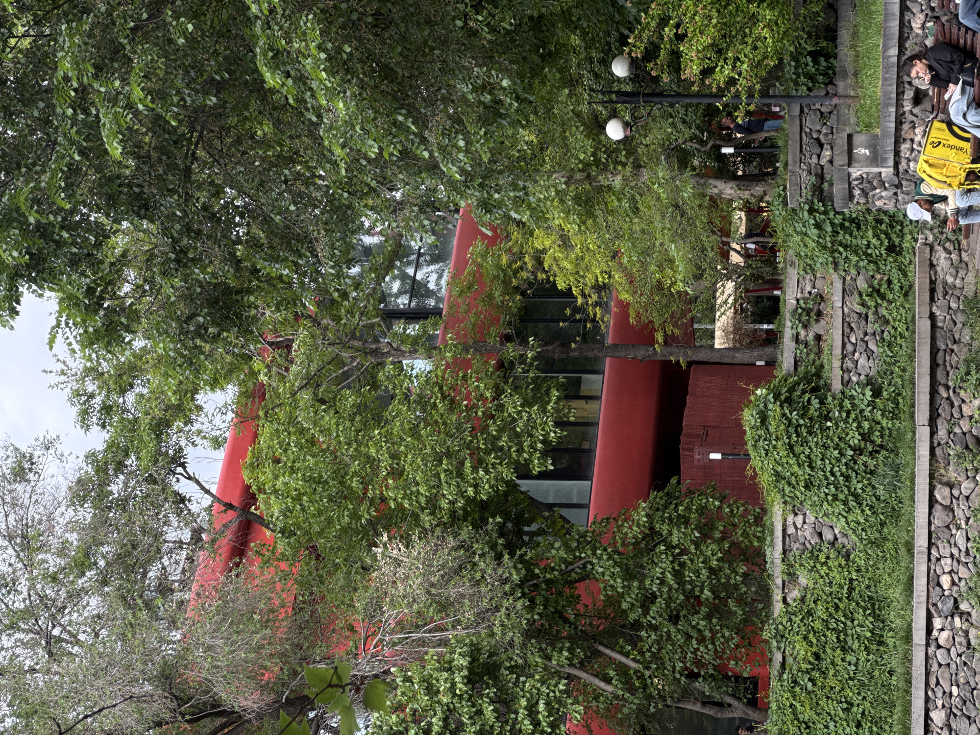 The image shows a red building with modern architecture, partially obscured by trees. The building has glass windows and red cylindrical columns. Next to the building is a walkway with stone and vegetation. There is a bench where two people are sleeping, one with a yellow Yandex.go bag. A lamp post is also visible. The image appears to be taken in a park setting in Yerevan, Armenia.