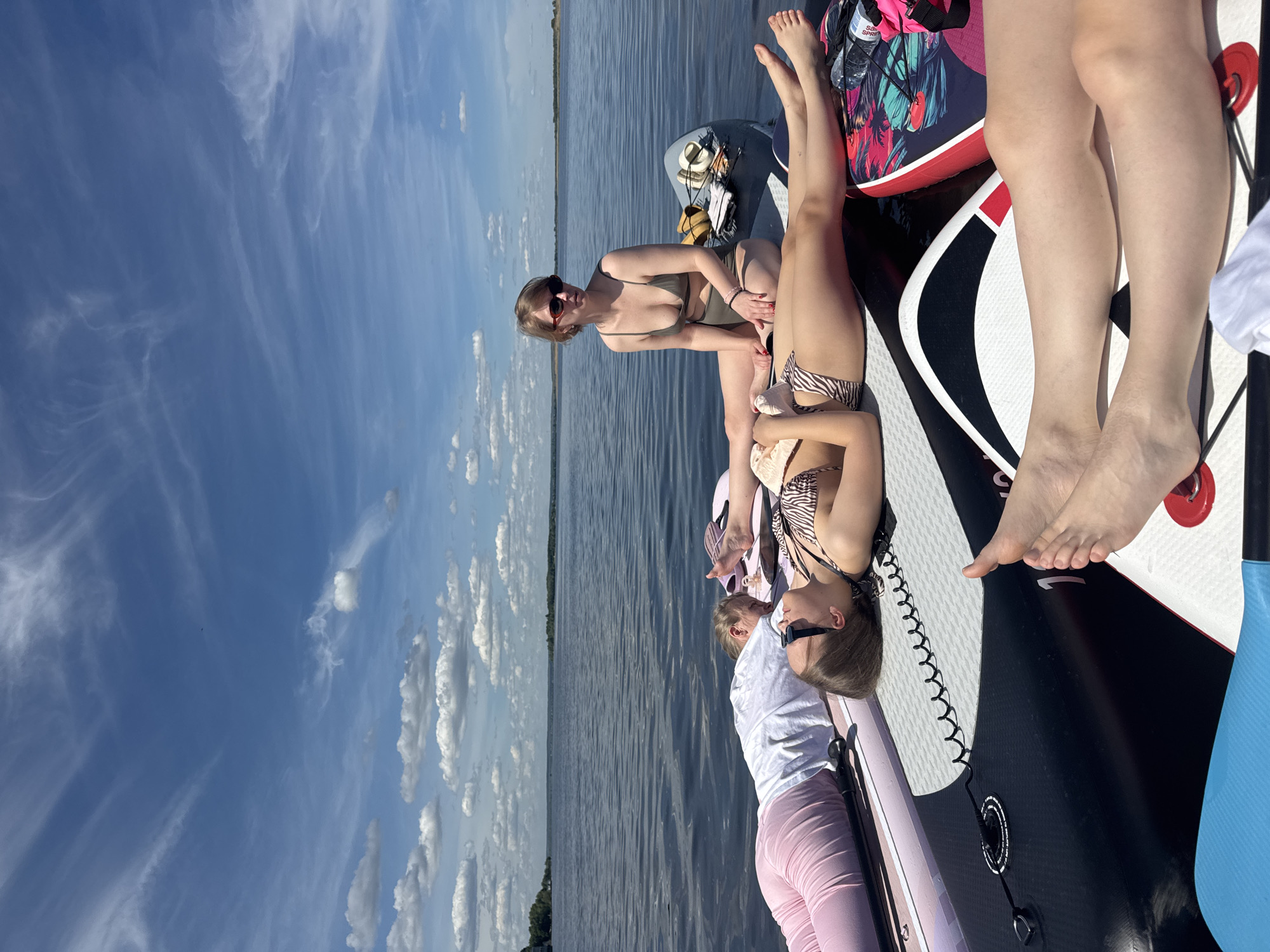 Four women are seen relaxing on paddleboards on a large, calm body of water under a bright blue sky with scattered clouds. The scene is set outdoors during what appears to be a sunny day, likely midday or early afternoon.
In the foreground and midground, three women are clearly visible. One woman in a green bikini and sunglasses is seated on a paddleboard. Another, wearing a zebra-print bikini, is lying down on a separate paddleboard. A third woman wears a white t-shirt, pink pants, and has sunglasses perched on her head, looking down. Portions of a fourth person's legs are also visible, suggesting more individuals are present.
The women are engaged in a leisurely activity, possibly sunbathing or enjoying a day out on the water. Various objects are scattered on the paddleboards, including a vibrant pink bag with a floral pattern, a water bottle with "SPRI" visible on its label, a straw hat, and yellow sandals. A blue paddle and a coiled paddle leash are also visible.
The surrounding environment features a wide expanse of calm, dark blue water reflecting the sky, with a distant, low-lying tree line indicating a shoreline. The sky transitions from mostly clear blue on the left to an area with more fluffy white cumulus clouds on the right, suggesting pleasant weather conditions ideal for outdoor water activities. The overall mood of the image is one of relaxation and enjoyment. The location context indicates this is likely in or near Усть-Курдюм, Russia.