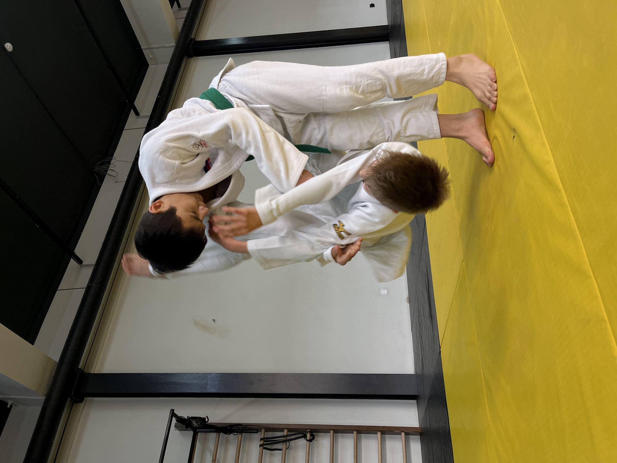 Two young males wearing white judo uniforms with green belts are grappling on a yellow mat. The location appears to be indoors in a gym, possibly in Krasnoyarsk, Russia, based on the uniform's national emblem. The background includes a black structural support and a white wall. The flooring is yellow. The males are actively engaged in a judo match, with one attempting to throw the other.