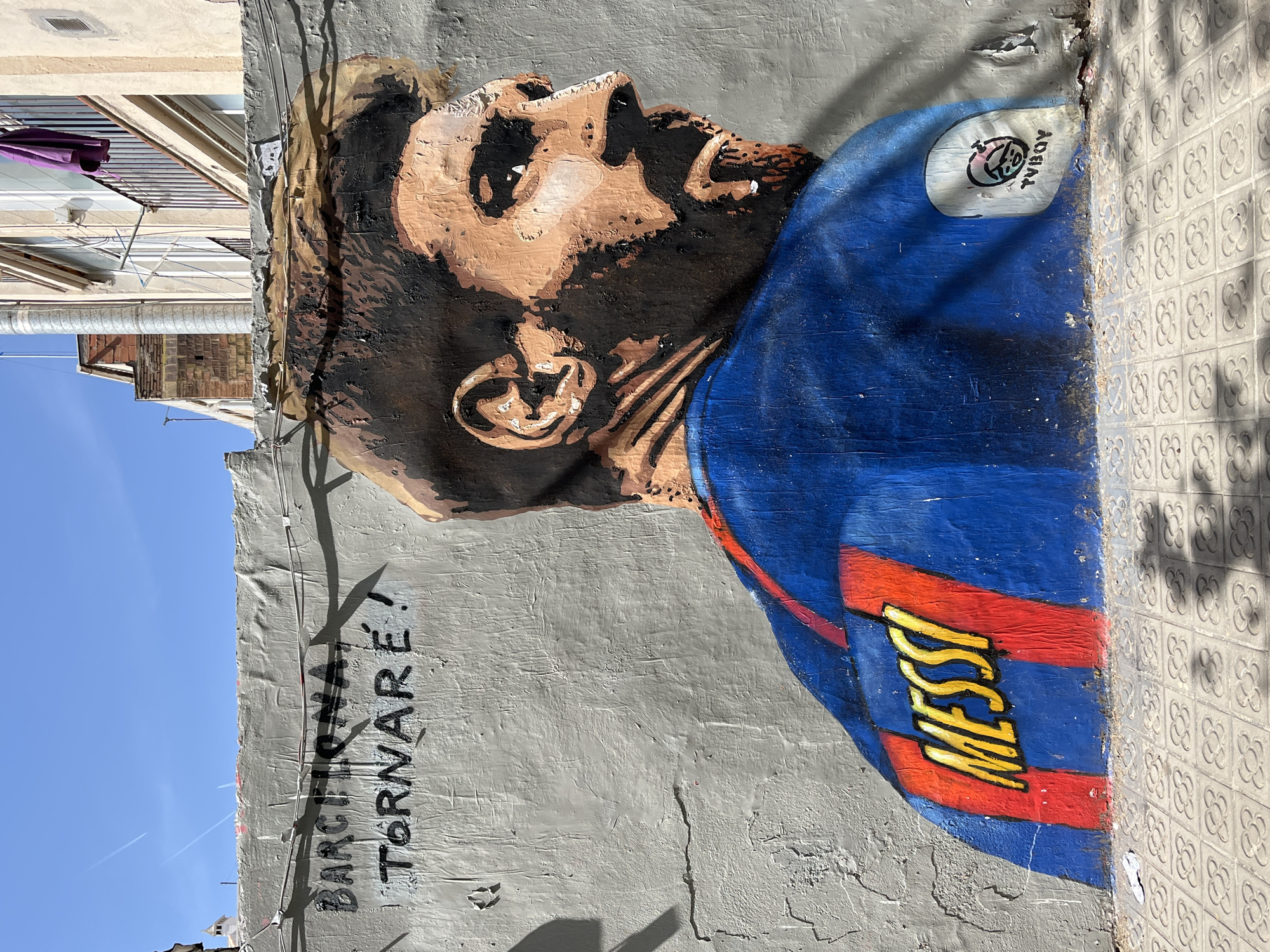 This media file features a vibrant street art mural of Lionel Messi painted on a textured grey wall, likely in Barcelona, Spain, given the location context. Messi is depicted in profile, looking upwards, wearing a blue and red FC Barcelona jersey with "MESSI" written in yellow on the lower back. The mural has a stylized appearance, with distinct brushstrokes forming his facial features and beard.
To the left of Messi's head, the artist's signature or logo, "TVBOY," is visible within a small cartoon face. Below Messi's portrait, on the grey wall, the Catalan phrase "BARÇA TORNARÉ!" is spray-painted in black, which translates to "Barça, I will return!".
The mural occupies the right side of the frame (when viewed in its upright orientation). The ground immediately next to the wall features characteristic patterned tiles, often seen in Barcelona. Above and to the left of the mural, parts of urban buildings are visible, including windows, balconies, and what appears to be a purple umbrella hanging from a line. The sky above is clear and bright blue, indicating good weather and strong sunlight, which casts distinct shadows on the wall and from the buildings. The time of day appears to be mid-day due to the high sun and strong shadows.