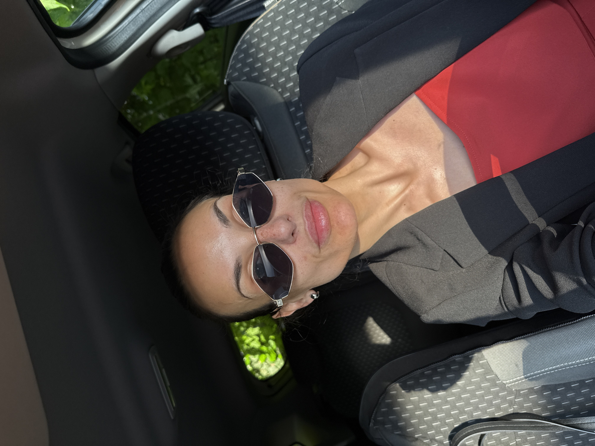 A woman is seated in a car. She is wearing a black blazer over a red top and sunglasses. The interior of the car is visible, including the seats and a glimpse of the outside through the window. The specific location within the city of Pleven, Bulgaria is not identifiable from the image.