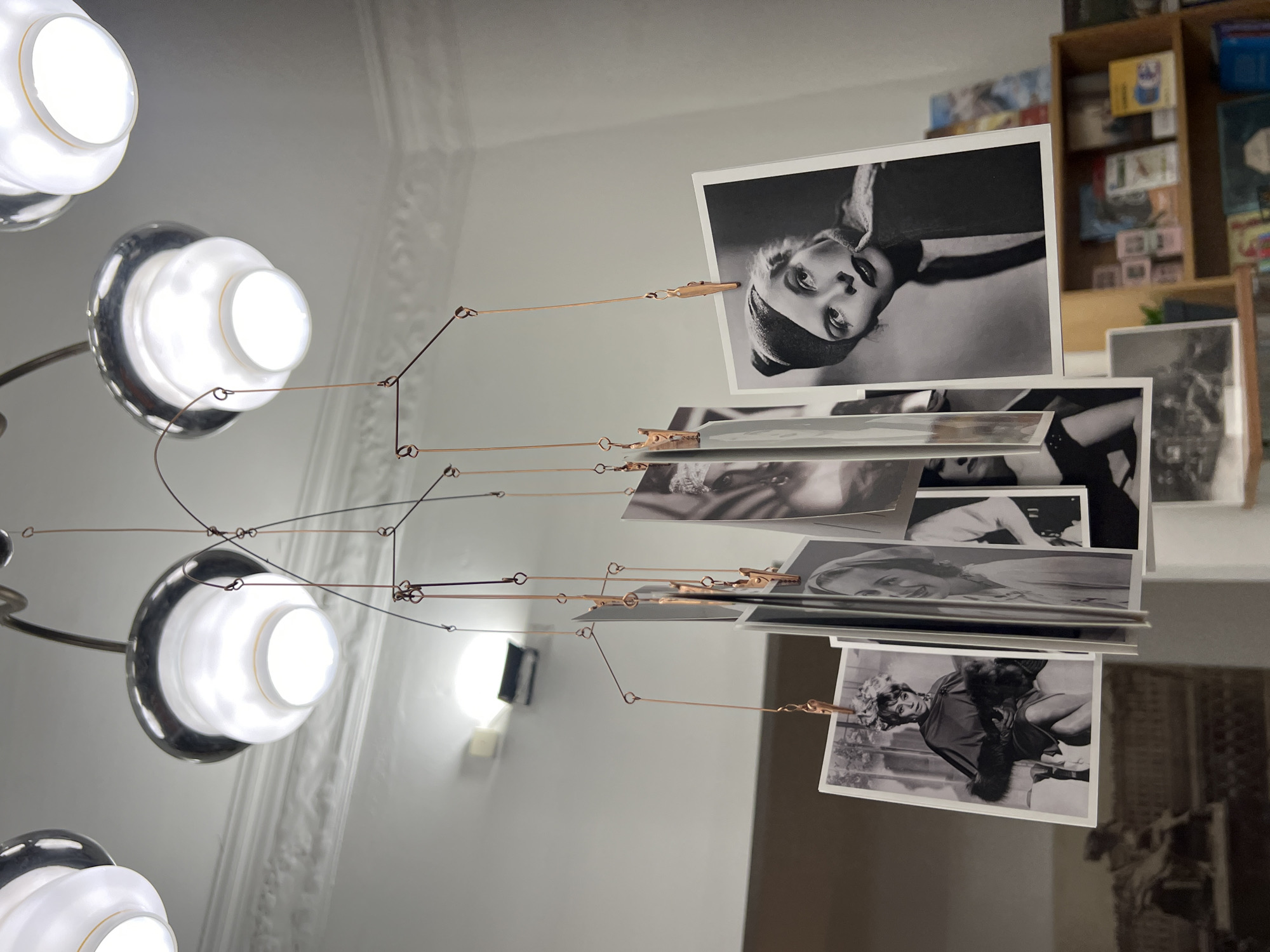 The image captures an artistic indoor display, showcasing a multi-tiered chandelier alongside a hanging mobile adorned with black and white photographs. The chandelier, situated on the left, features several light sources, each with a rounded, white frosted glass shade nested within a reflective chrome or silver base.
A delicate mobile system, suspended from the ceiling, is fashioned from thin, copper-colored wires. These wires crisscross and extend downwards, supporting small metallic clips. Attached to these clips are numerous black and white portrait photographs, primarily featuring women in what appears to be vintage fashion, evoking classic cinema or fashion photography. A prominent photograph in the upper right shows a woman with striking eyes and a dark head covering, gazing upwards. Another visible photo depicts a woman in a fur stole or coat. The layered arrangement of the photos creates a dynamic visual display.
In the background, a light-colored wall exhibits intricate, ornate molding near the ceiling, characteristic of classical European architecture, aligning with the Saint Petersburg location context. Further right, a wooden bookshelf is visible, filled with various items such as books and small boxes, adding to a curated or lived-in atmosphere. The interior is brightly lit, suggesting either ample daylight or strong artificial illumination. No discernible text is visible on the photographs or within the surrounding environment. The scene overall conveys an aesthetic of artistic appreciation and vintage elegance within a sophisticated setting.
