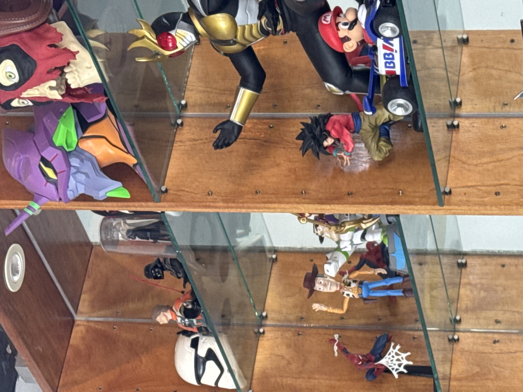 The image displays a wooden display shelf filled with various collectible figurines. On the top shelf, from left to right, there is a white stormtrooper helmet, a figure in an orange and white pilot suit, a Darth Vader figure with a red lightsaber, and a purple and green Evangelion head alongside a skull-like mask with red accents. The middle shelf features a large figure in a black and gold suit with a red helmet, a smaller figure of Goku in a red top and brown pants, and a blue race car with a Mario figure driving it. The bottom shelf shows a Spiderman figure in a dynamic pose with a web, a Woody figure from Toy Story, and what appears to be a Buzz Lightyear figure. The background is a light-colored wall. The scene is indoors, with the lighting suggesting it might be daytime or under artificial lights. The figurines are arranged on glass shelves supported by metal pins.