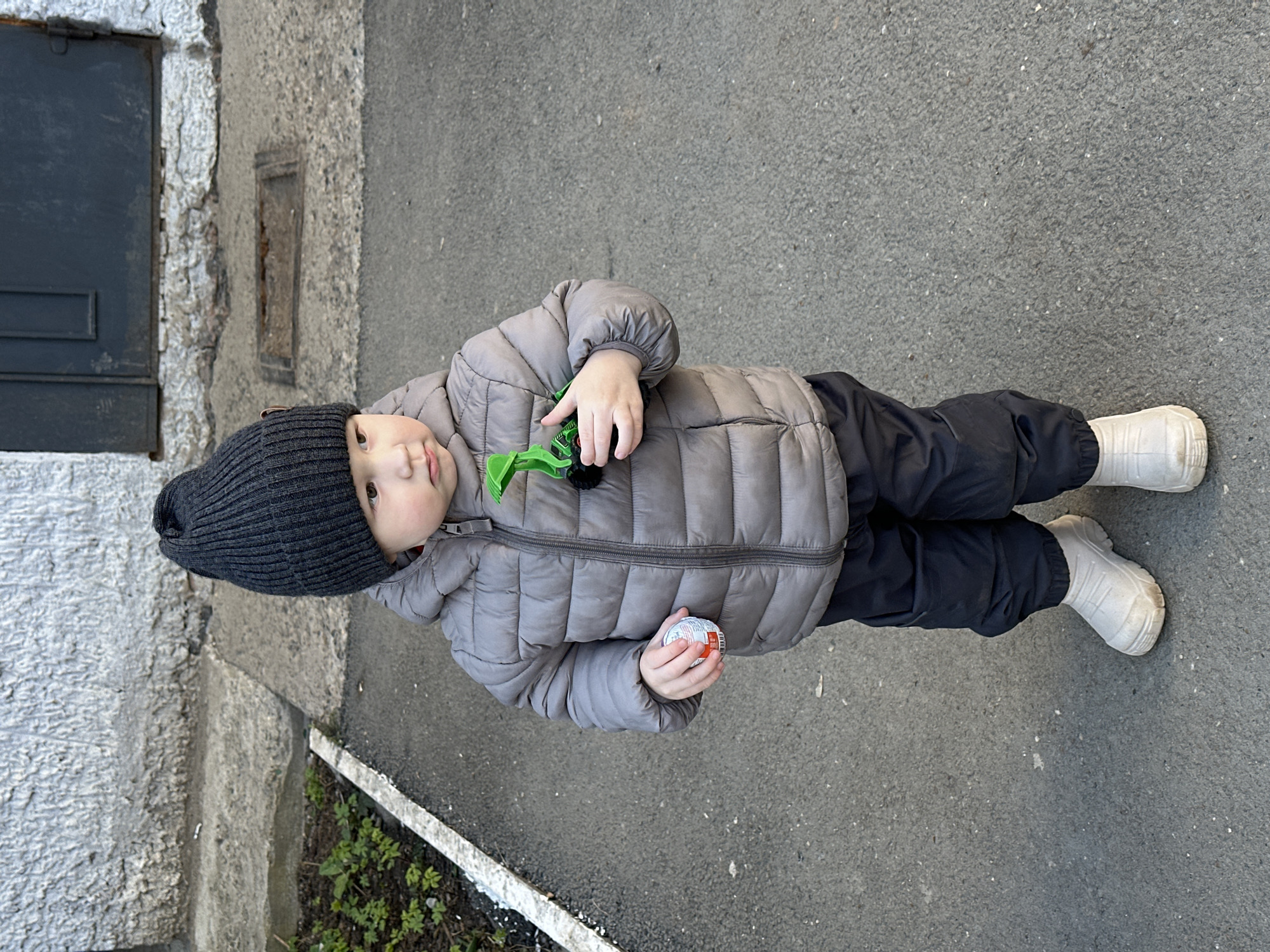 A young child, likely a toddler, is standing on a paved surface, possibly a sidewalk, adjacent to a building. The child is wearing a dark gray knit hat, a light gray quilted jacket, dark pants, and white boots. The child holds a small, round object in one hand and a green plastic toy in the other. The building beside the child has a gray exterior and includes a door with a dark gray inset panel and a smaller inset panel above it. Green vegetation is visible along the base of the building. The location is Votkinsk, Russia.