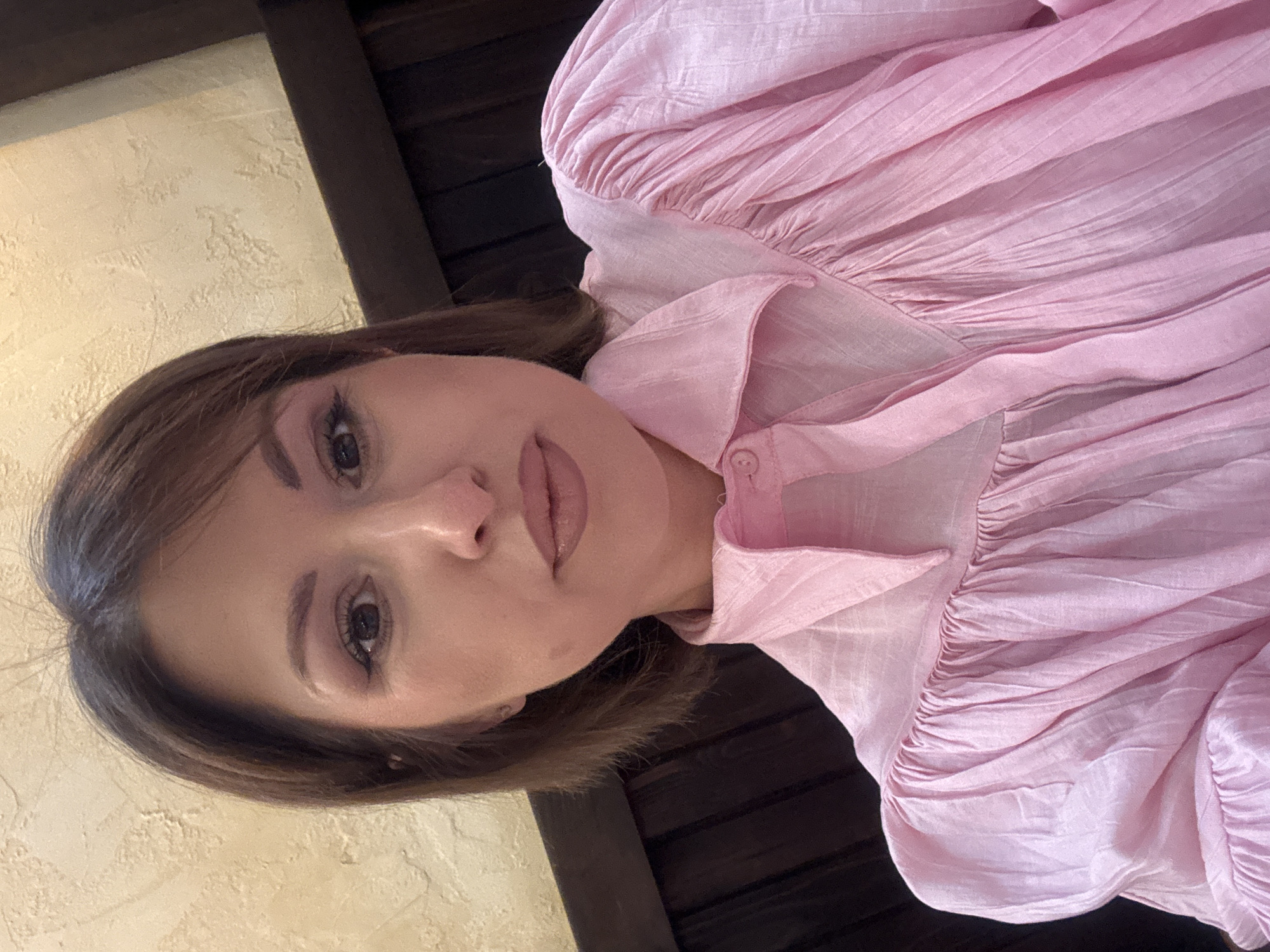 A medium, close-up shot shows a woman wearing a pink blouse. The background shows a cream-colored wall and dark wooden beams, indicating an indoor setting that could be located in Elin Pelin, Bulgaria or elsewhere. The woman's head is slightly tilted. The image is oriented sideways.
