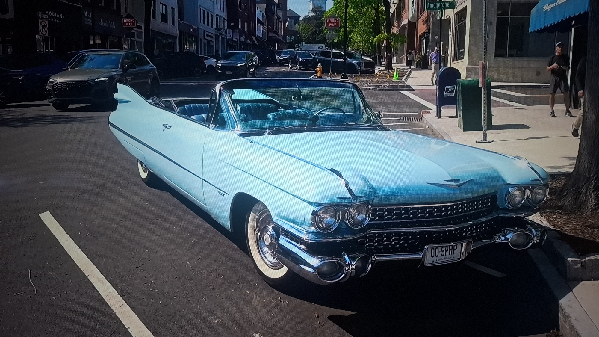 The image depicts a light blue vintage Cadillac convertible parked on a city street. The car is the primary subject, showcasing its distinctive fins and chrome details. A license plate reading "00-SPHP" is visible on the front. The surrounding environment indicates an urban setting, with buildings lining the street. Other vehicles, including a dark-colored SUV and several cars, are parked or moving in the background. Sidewalks and pedestrian crossings are present, with a few individuals walking in the distance, some near a blue mailbox and a street sign indicating "Amogerone". A "Wrong Way" sign is also visible, and there are other street signs mounted on buildings. A tree is present on the sidewalk next to the car. The scene takes place during daylight, with moderate sunlight casting shadows. The city and state are unspecified.
