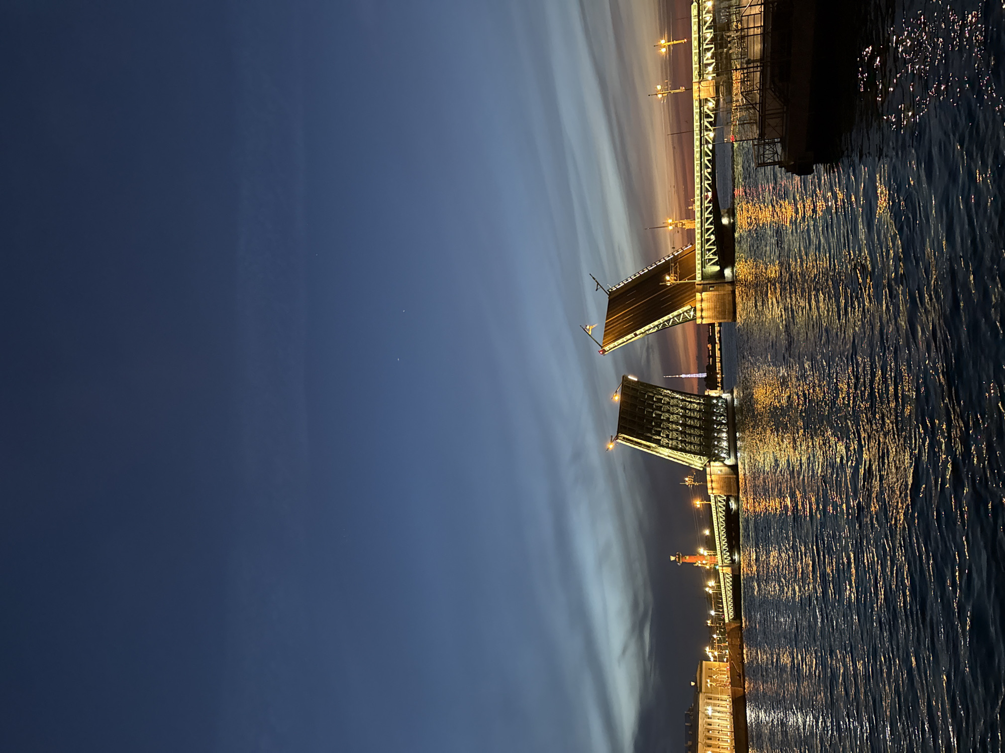 The image displays the illuminated Palace Bridge (Dvortsovy Most) in Saint Petersburg, Russia, at dusk or night, with its central spans raised. The drawbridge is angled upwards, with its internal structure visible and its edges and surface brightly lit by warm yellow artificial lights. These lights cast prominent, shimmering reflections on the dark, rippling surface of the Neva River, which occupies the lower half of the frame.
On the left bank, a section of a large, classically styled building is visible, also illuminated. To the right of this building, one of the Rostral Columns stands prominently, glowing with an orange light. In the background, between the raised sections of the bridge, a tall, slender spire, likely the Peter and Paul Cathedral or the Saint Petersburg TV Tower, is faintly visible.
The sky above is a gradient of deep blue at the top, transitioning to lighter blues and faint hints of orange and pink near the horizon, indicating residual twilight. Wispy cloud formations stretch across the sky. Two minute, bright points, possibly stars or distant lights, are discernable in the upper left portion of the sky. In the lower right foreground, a dark, indistinct structure resembling a boat or barge is partially visible. No identifiable people are present in the scene.