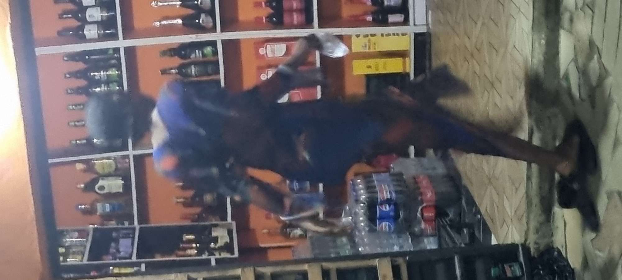 A person stands in front of a store shelf in what appears to be a liquor store, likely in Lagos, Nigeria. The shelves behind them are filled with bottles of liquor and other beverages. The person is wearing sandals, shorts, and a shirt. They hold a water bottle in their left hand. A shelf below contains water bottles. The background shows a patterned curtain.