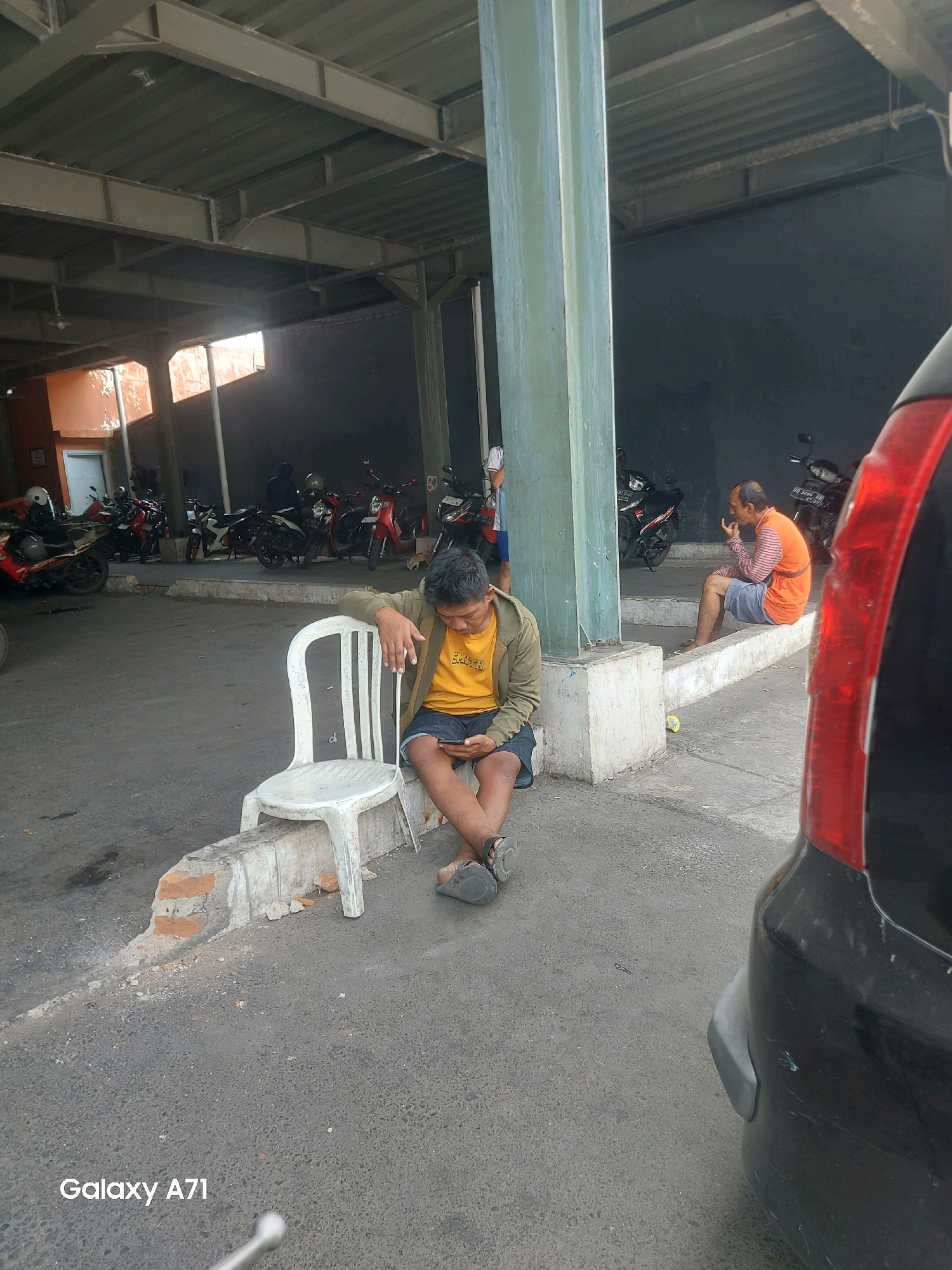 The image depicts an outdoor covered area, likely a parking lot or waiting area, with a concrete floor and a corrugated metal roof supported by pillars. Several motorcycles are parked in the background. In the foreground, a young man is seated on a white plastic chair, looking at his phone. He is wearing a yellow t-shirt and dark shorts. To his right, another man is sitting on a raised concrete ledge, wearing an orange and striped shirt and shorts. The overall scene suggests a casual, possibly waiting, environment within the city of Makassar, Indonesia. The presence of the "Galaxy A71" watermark indicates the image was taken with a Samsung Galaxy A71 smartphone.