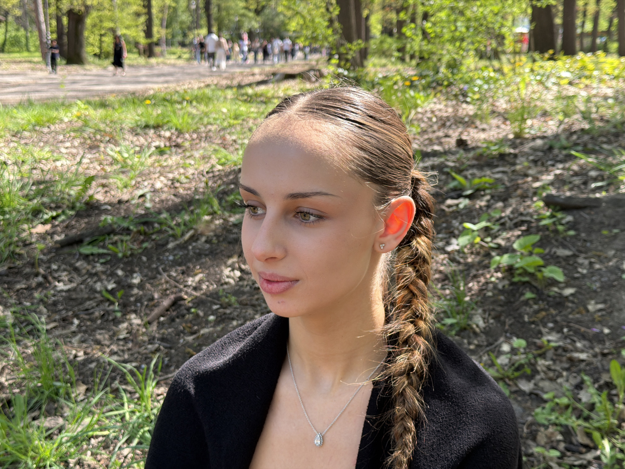 A young woman with light skin and brown hair braided and pulled back from her face is seated outdoors, likely in a park or wooded area. She is wearing a black sweater and a silver necklace with a pendant. In the background, there is a path with several people walking along it, and there are trees and green foliage surrounding the area. The scene is likely in a park setting in a city, given the presence of both nature and a visible pathway with people. The location of the image is most likely Sofia, Bulgaria, based on visual clues and the information provided.