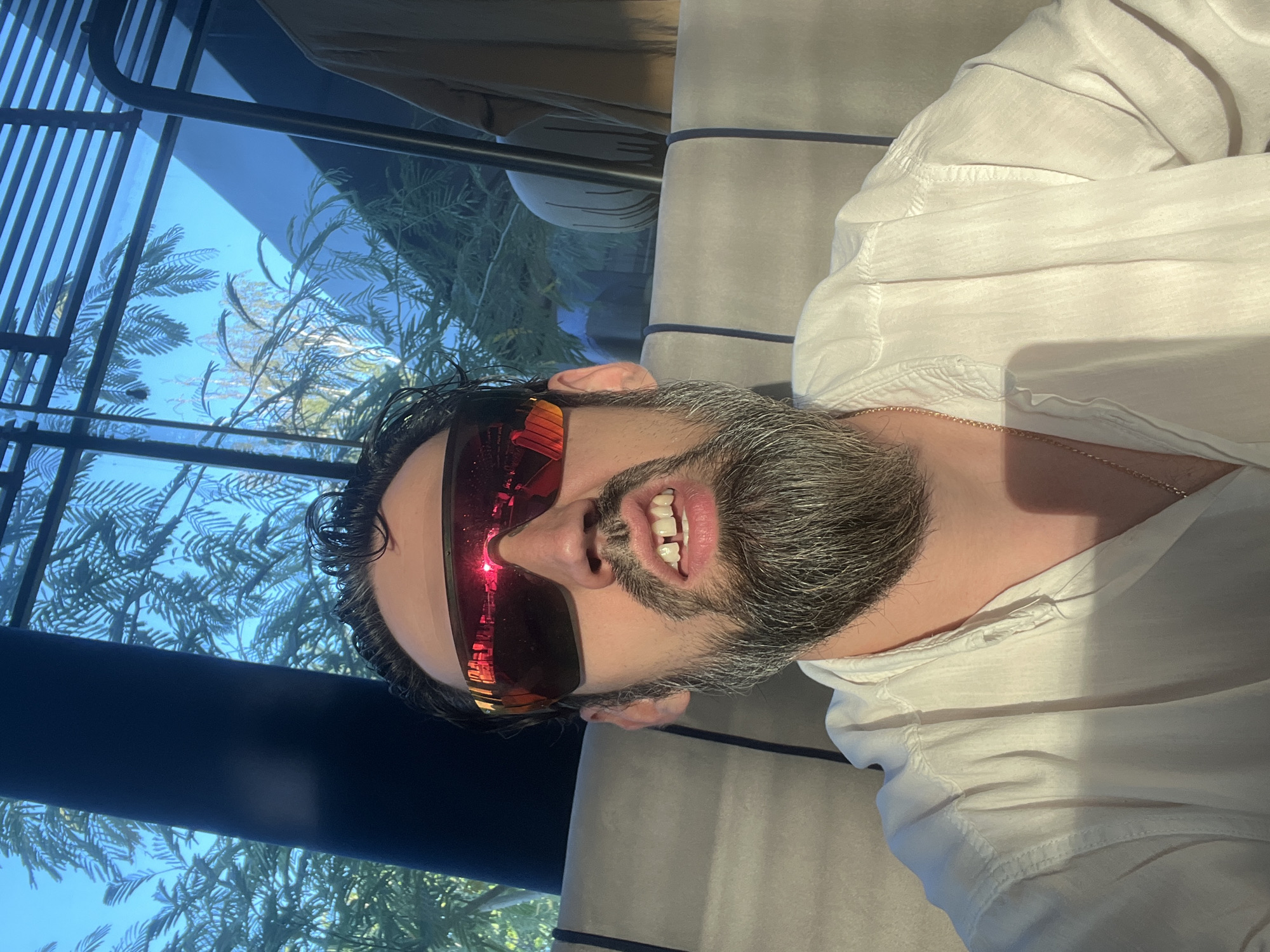 A man with a grey beard and a white shirt is seen wearing red mirrored sunglasses. He is sitting in what appears to be an indoor setting with a window showing greenery and a blue sky. The man's mouth is slightly open, revealing a gap between his front teeth. The lighting suggests it is daytime. The scene is in Tiraspol, Moldova.