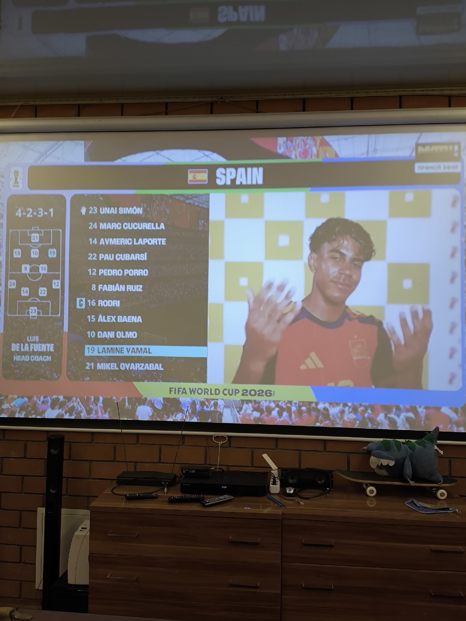 The image shows a projection screen displaying a football team's lineup and a player. The text on the screen indicates it is the FIFA World Cup 2026. The lineup for Spain is shown, with numbers and names of players such as "23 UNAI SIMON," "24 MARC CUCURELLA," and "19 LAMINE YAMAL" highlighted. A young man in a red and yellow football jersey is prominently featured on the right side of the screen, with his hands raised. Below the projection screen, a wooden dresser is visible with various electronic devices, including a soundbar, a DVD player, a satellite receiver, and remote controls. A skateboard with a plush toy is placed on top of the dresser. A tall black speaker stands to the left of the dresser. The background appears to be a brick wall. The scene is indoors. The location in the city of Pokrov, Russia is unknown.