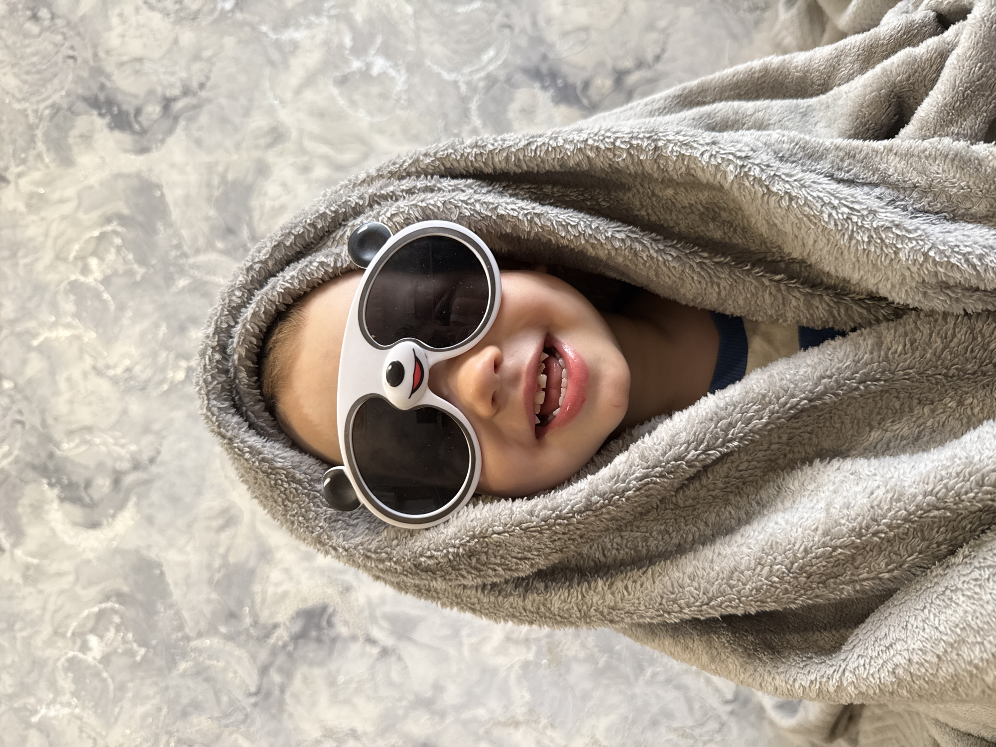 A young child is swathed in a grey, plush blanket and is lying on a surface with a grey and white mottled pattern, possibly a bed or floor. The child is wearing white panda-shaped sunglasses, revealing only the bottom half of the face. A smile reveals some missing front teeth. The surrounding area is indistinct, but suggests an indoor setting. Location: Unknown, Unknown.