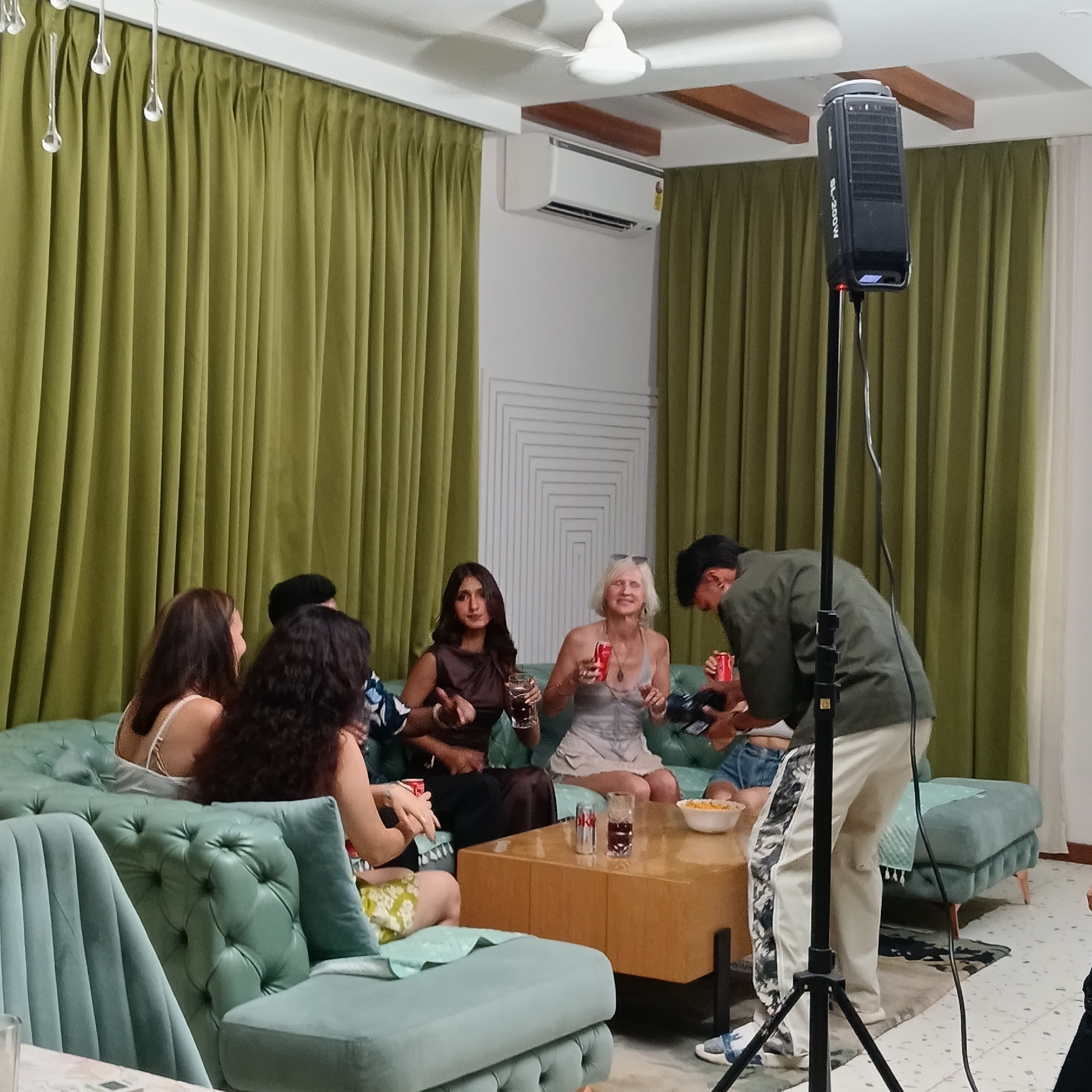 The image depicts a group of people in an indoor setting, likely a living room, in Calangute, India. Five individuals are seated on a tufted, light-green sofa. They are holding beverages in cans and glasses. A man standing to the right of the group appears to be interacting with them. A wooden coffee table is in front of the sofa, with a bowl of snacks and beverage containers on it. A large, black audio speaker mounted on a tripod is positioned to the right of the group. The room is decorated with green curtains, a white air conditioning unit, and a geometric wall design. Two tufted, light-green chairs are visible in the foreground. The ceiling has a white ceiling fan and wooden beams.