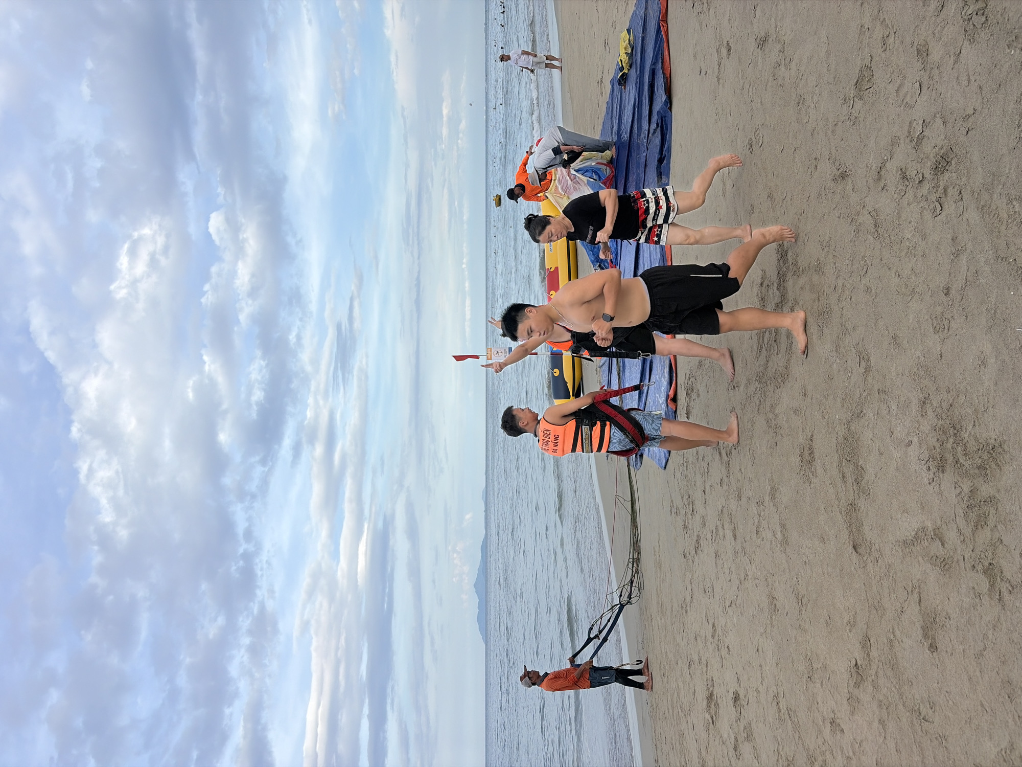 The image depicts a beach scene in Đà Nẵng, Vietnam. Several individuals are gathered near the waterline, with the ocean and a distant island visible in the background under a cloudy sky. In the foreground, a shirtless young man is running towards the right of the frame, wearing black shorts. To his left, a woman with dark hair is also running, dressed in a black t-shirt and striped shorts. Behind them, a person in an orange life vest, identified with text on the back, is interacting with a yellow inflatable watercraft. Another individual in an orange shirt is holding onto ropes connected to some equipment on the sand. Further in the background, other people are seen on the beach and in the water, suggesting a public recreational area. The sand is light brown, and a large blue tarp is spread out on the beach.