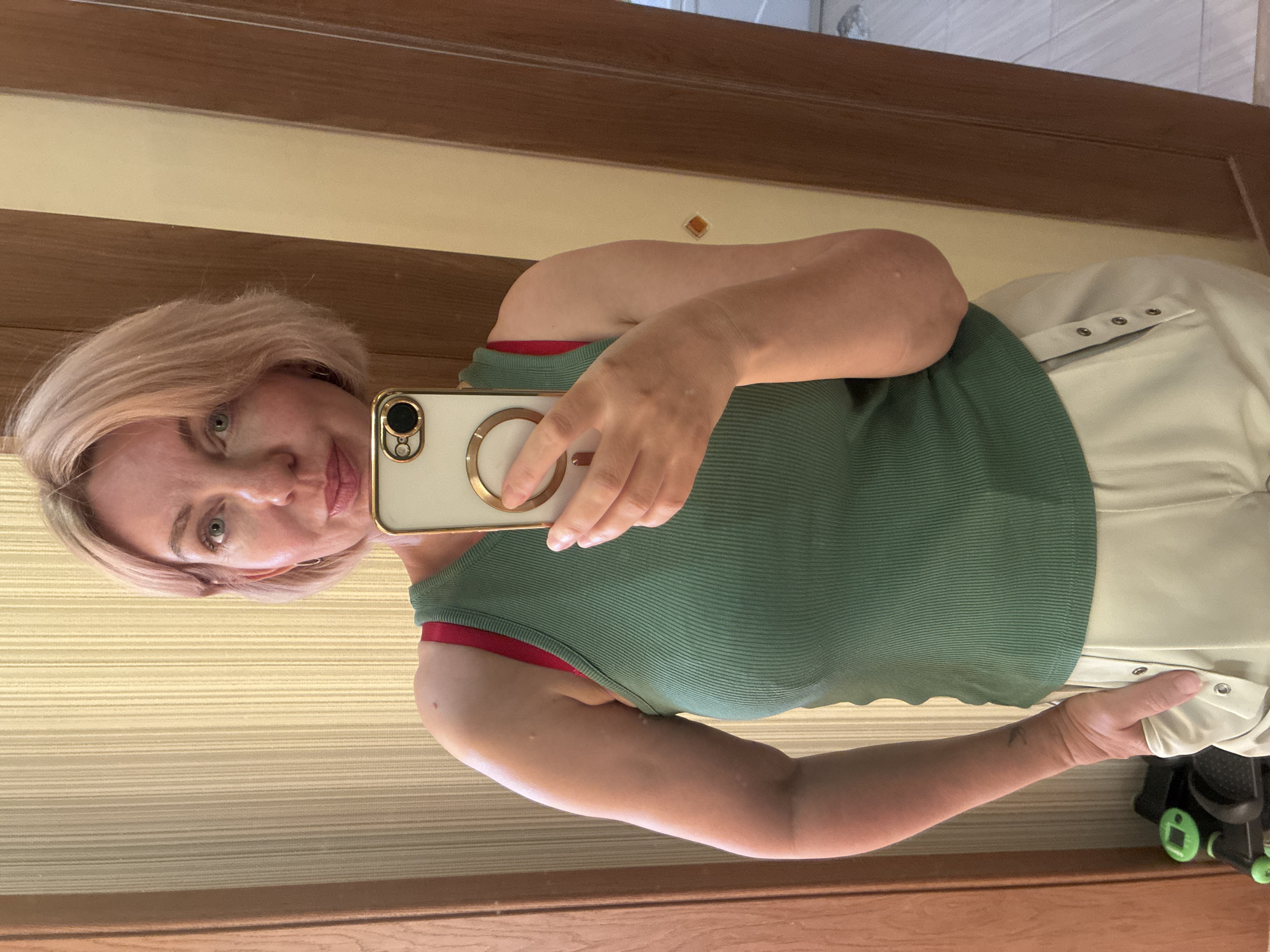 A woman, appearing to be in her 40s or 50s, with short, light pinkish-blonde hair and light-colored eyes, takes a mirror selfie. She has a slight smile. A small mole is visible on her upper left arm, and multiple small skin tags are present on both upper arms. A small, dark bird tattoo is visible on her left inner wrist. She wears a ribbed, dark green sleeveless tank top, revealing a red bra strap on her left shoulder. She is also wearing light-colored, possibly beige or off-white, pants with decorative snap buttons along the outer seams of the pockets. Her left hand is inserted into her left pant pocket, while her right hand holds a smartphone, equipped with a light-colored case and a gold-toned magnetic ring holder, positioned just below her chin. The background consists of a light-colored, vertically striped wallpaper. Dark brown wooden door frames are visible on both the left and right sides of the image. A section of a light-colored tiled floor is visible on the lower right. On the lower left, a dark object with green wheels, possibly a child's toy, is partially visible on the floor. The exact location beyond an indoor setting cannot be determined from the visual evidence.