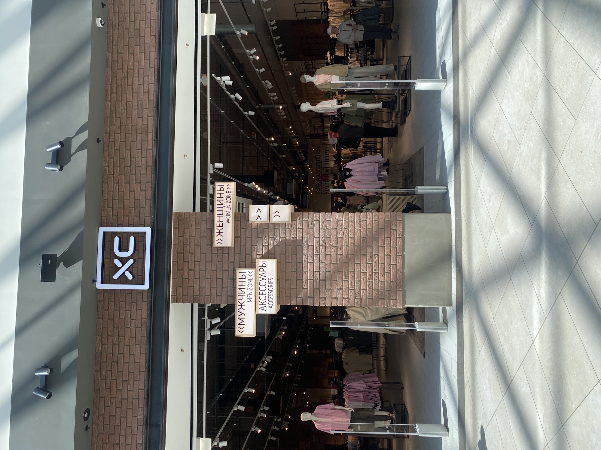 The image captures the entrance to a clothing store located in Perm, Russia. The store entrance features brickwork on its façade with signs indicating "Women Zone," "Men Zone," and "Accessories" in both Russian and English. Above the entrance is a white illuminated sign with a logo. Mannequins dressed in clothing are visible inside the store, which is well-lit. The interior features a long hallway with clothing racks, typical of a department store. The photograph is taken during the day, with natural light coming through the large windows. The floor outside the store is tiled with a grid pattern. The phrase "KAMSKY" can be seen inside the store.