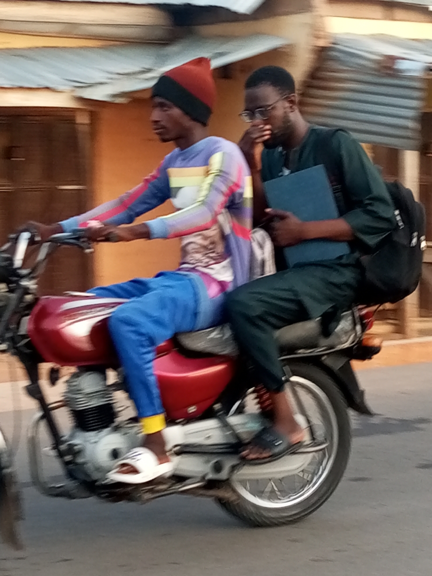 Two men are riding a red motorcycle on a street in an urban environment. The driver is wearing a red and black hat, a patterned long-sleeved shirt, and blue pants. He is wearing white sandals. The passenger is wearing glasses, a dark green shirt, and dark pants. He is carrying a blue book and a black backpack. The buildings in the background are nondescript and made of corrugated metal and painted cinder blocks. The location is the city of Unknown, Unknown.