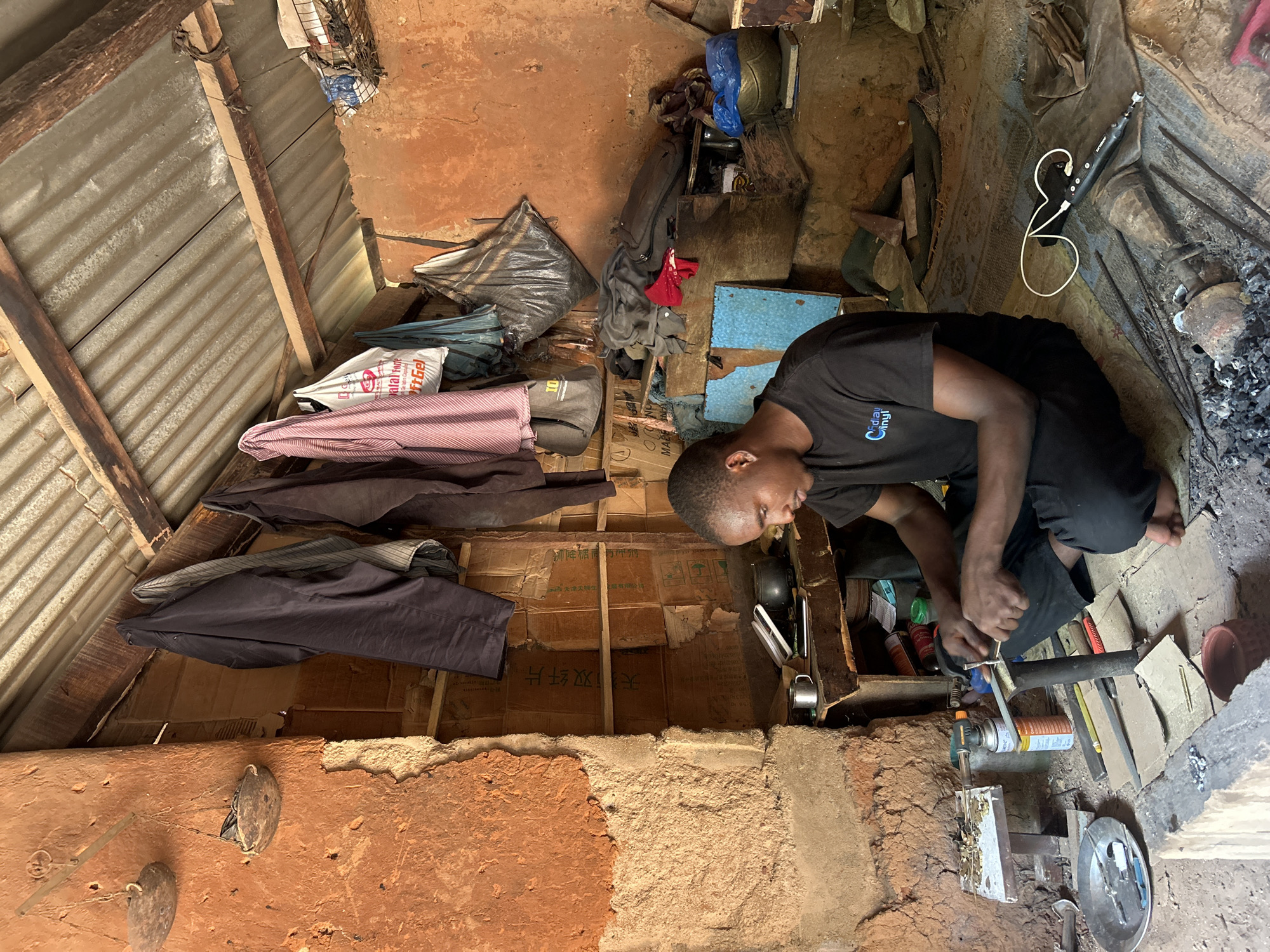 A young Black man is working in what appears to be a small workshop or home in Unguwar Shanu, Nigeria. He is seated on the floor, focused on a metal object held in a clamp on his workbench. He is wearing a black t-shirt with a blue and white logo that reads "Cadillac Vinyl." His legs are crossed and he is wearing black pants.
The workshop is rustic, with walls made of reddish-brown mud or clay. The ceiling is made of corrugated metal sheeting supported by wooden beams. Various items are stored around the workshop, including what appear to be tools, clothing hanging from hooks, sacks, and bags. On the workbench, there are various tools, including what looks like a soldering torch, metal files, and small metal pieces. A metal plate sits on the floor to his left with several tools on it. In front of him, a small charcoal brazier is visible with some metal rods and tools scattered around it. The floor is covered with a patterned rug and dirt.
The overall impression is of a working environment that is functional but simple, likely typical of a craftsperson's space in this region. The lighting suggests it is daytime, with light filtering in from the open front of the structure.