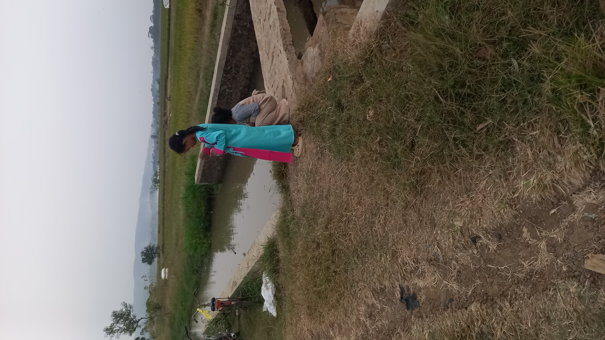 In Sidamulya, Indonesia, on a cloudy day, two young girls are near a narrow, muddy canal. The girl in the foreground, facing right, wears a turquoise dress with a pink trim and light-colored clogs. She is looking down at something in her hands. Behind her, a second girl crouches, wearing a plaid and beige outfit. The canal runs parallel to a path made of concrete and dirt, bordered by dry grass and some vegetation. In the background, a lush green rice field extends towards a hazy mountain range under an overcast sky. A bicycle with a yellow bag is visible to the left, and a white sack lies on the grass near the canal.