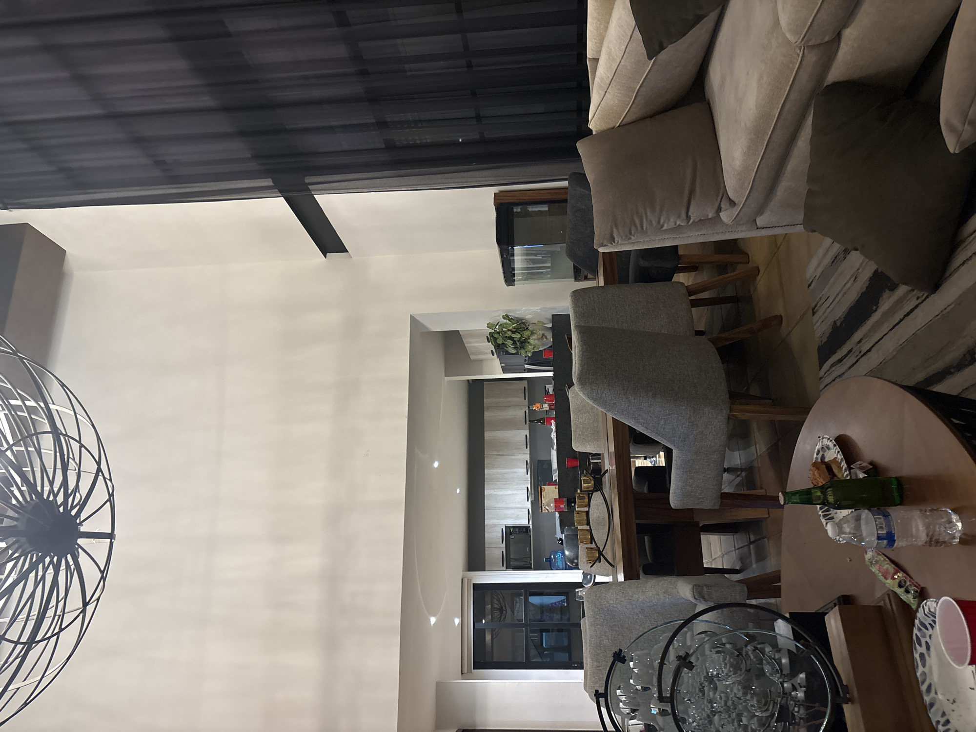 The image depicts an interior setting, likely a residential dining and living area. The space includes a light-colored couch with a dark pillow, and dining chairs. There is a dining table with beverages and other items. In the background, a kitchen area is visible. The lighting suggests an indoor environment with possible natural light. The presence of common household items indicates a domestic setting. The image does not provide enough information to determine if it is located in Santa Cruz Tehuixpango, Mexico. No people are visible.