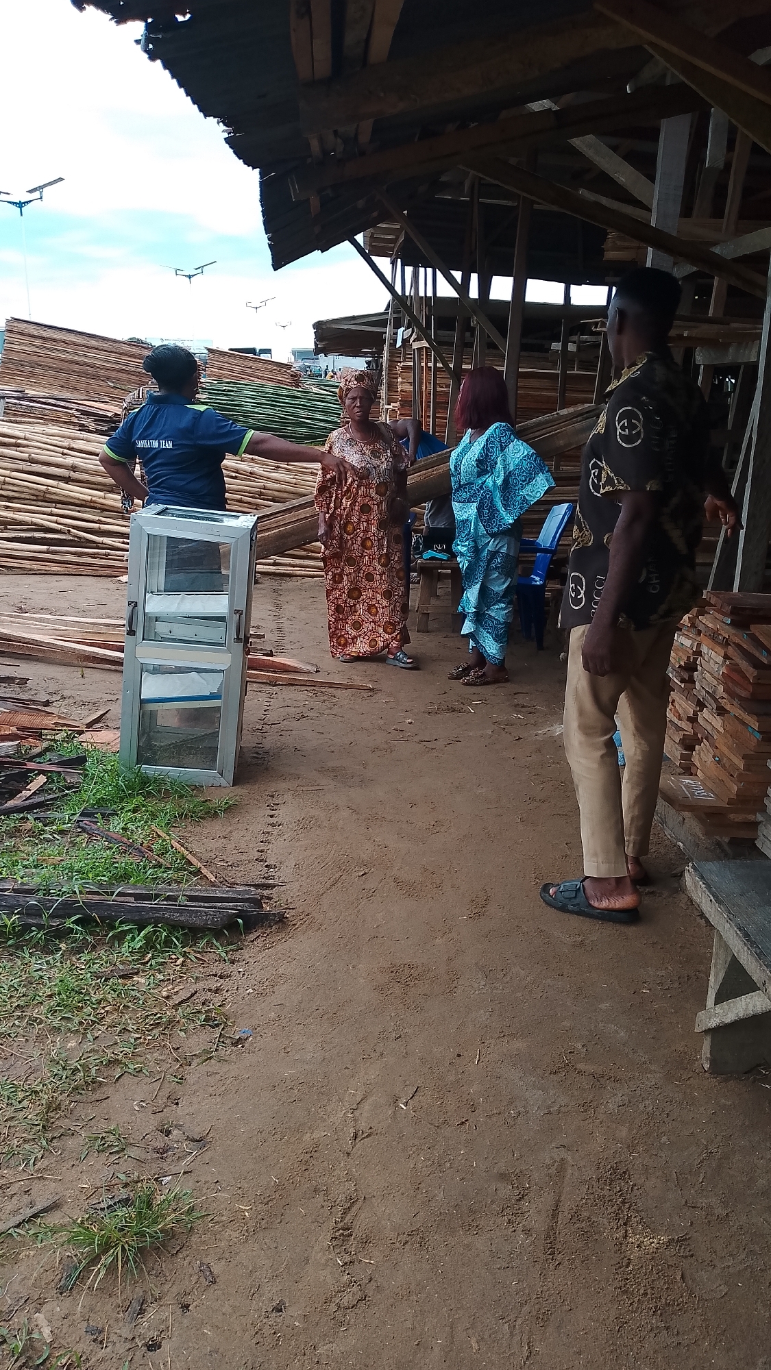 A medium-shot image captures a scene in Asubi, Nigeria, possibly a lumber or construction site. The ground is earthen, with scattered vegetation and debris. A stack of bamboo and lumber is visible in the background, partially covered by a makeshift wooden structure. Five people are present: one in a dark blue shirt marked "SANITATION TEAM," gesturing with their right arm; a woman in a brown and orange patterned dress and headwrap; a woman in a blue patterned dress; a man in khaki pants, a dark patterned shirt, and sandals, standing with his back mostly to the camera; and another woman that is partly behind the first woman. To the left of the "SANITATION TEAM" member is a small, free-standing aluminum and glass cabinet. Beside the man is a stack of wood.