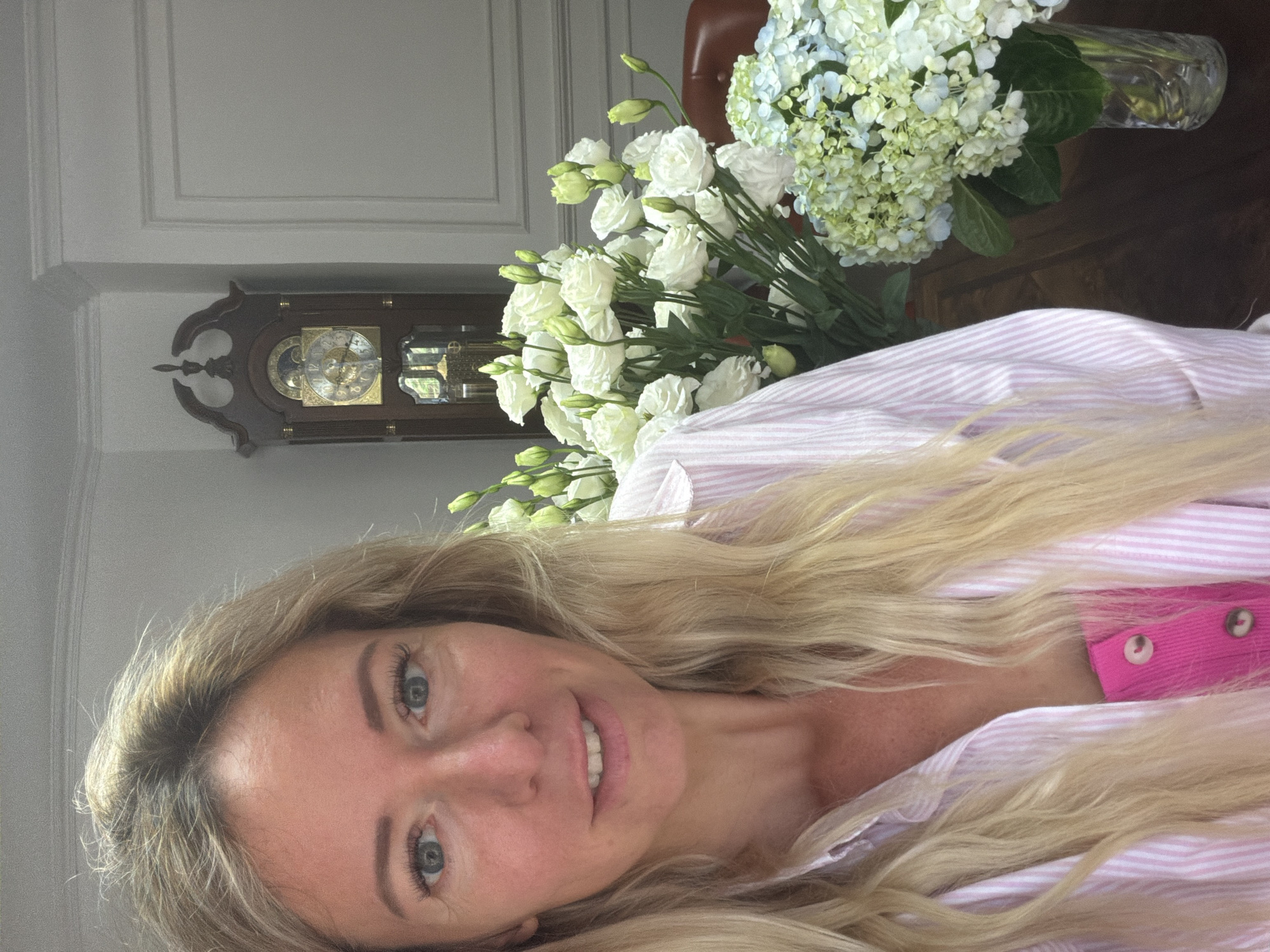 A woman with long blonde hair and blue eyes is taking a selfie in a room. She is wearing a pink and white striped shirt over a bright pink tank top. To her right are two vases of flowers, one with white roses and the other with blue and green hydrangeas. In the background, a grandfather clock stands against a wall. The setting appears to be indoors, possibly a home or a formal dining area. There is no indication that this scene is located in Xuân Trường - Đà Lạt, Vietnam.