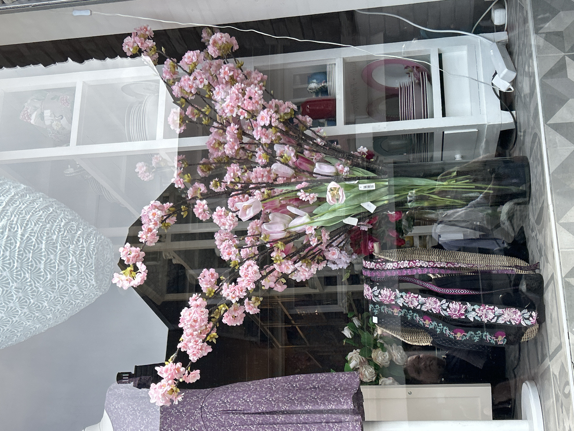 The image depicts a store window display in Waldbröl, Germany. In the foreground, a large arrangement of artificial pink cherry blossoms and pink tulips in a tall, dark glass vase dominates the center of the frame. To the left, a mannequin is dressed in a purple floral print dress. Behind the flowers, a white shelving unit displays stacks of plates and decorative items. The reflection in the window shows a building with a dark roof and a chimney. Below the flowers, a wicker basket holds several decorative fabric trims with floral embroidery. A person's face is visible in reflection behind the basket, looking out from within the store. The flooring is tiled with a geometric pattern. The overall scene suggests a retail environment, likely a boutique or gift shop.