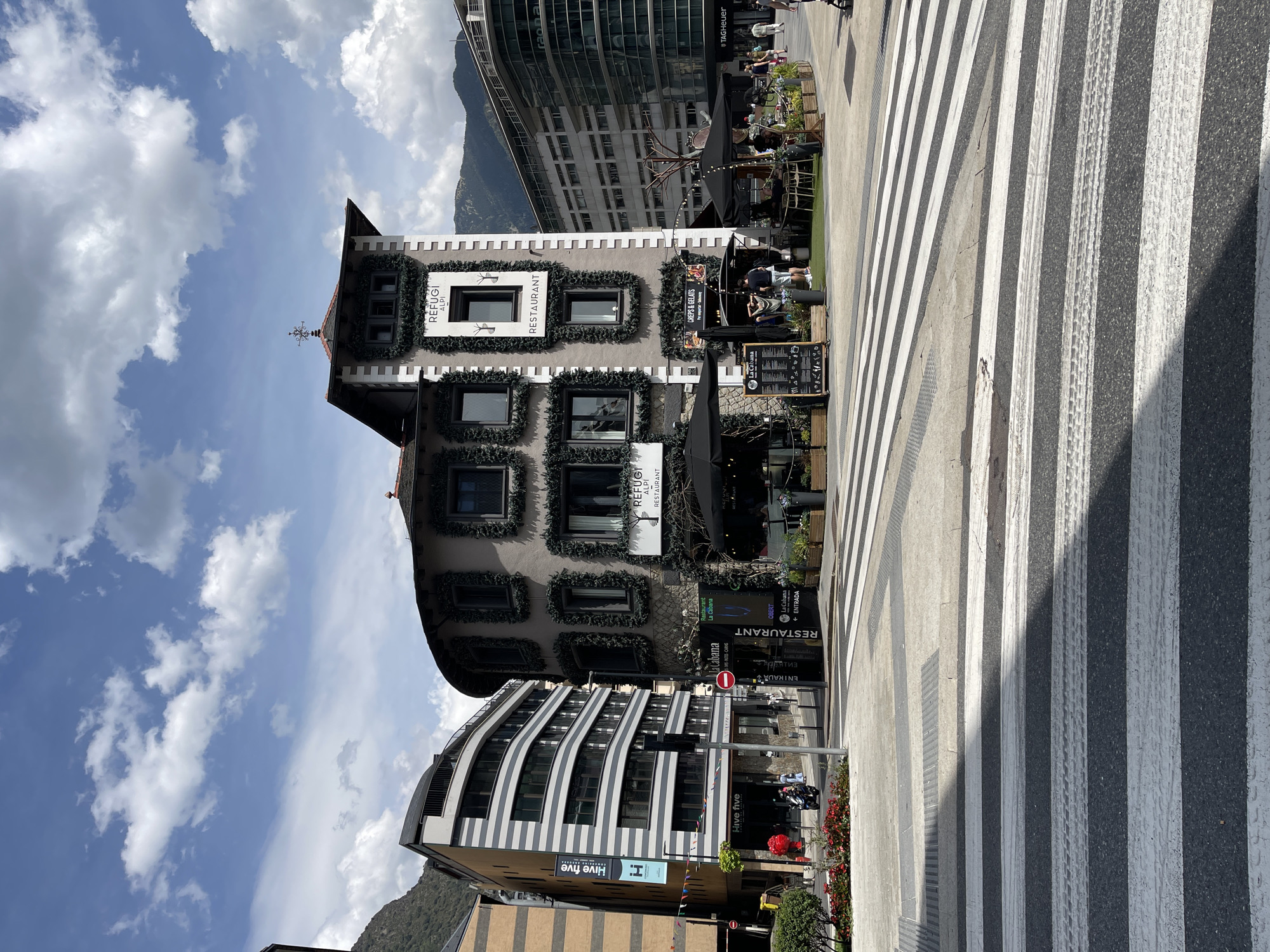 This is an eye-level, medium shot of a city street with a pedestrian crossing in the foreground and a curved building with a tower to the right of center. The building is decorated with dark green garlands around its windows and has signs for "REFUGI ALPI RESTAURANT". The street has a crosswalk with black and white stripes, some of which are marked by tire tracks. To the left of the curved building is a modern, multi-story building with horizontal black and white stripes and a sign for "Hive five". In the background, there are more modern buildings and mountains. Several people are visible walking and sitting at outdoor tables in front of the restaurants. The sky is blue with scattered white clouds, and the lighting suggests it is daytime.
