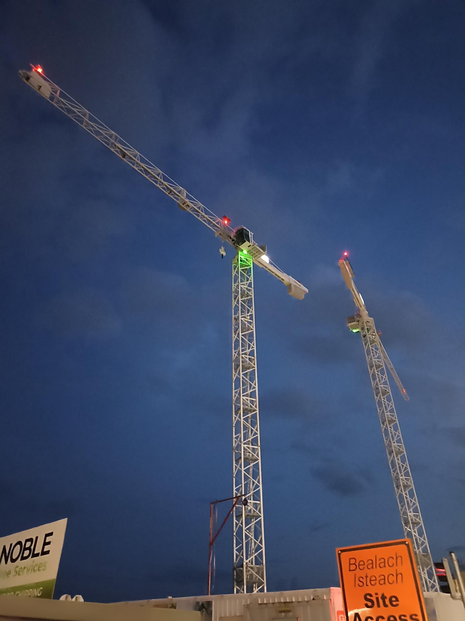 The image shows two construction tower cranes against a dark blue, cloudy sky. The crane on the left is closer to the camera and its boom is angled upwards and to the right. It has red lights at the tip and a green light on the operator's cab. The crane on the right is further away and its boom is angled upwards and to the left. It also has red lights at the tip and a green light on the operator's cab.
In the foreground, a white sign with black lettering reads "NOBLE" and below it "Tree Services". To the right of this sign, a bright orange sign with black text is visible. The text on the orange sign is in both Irish and English: "Bealach Isteach Site Access". This sign indicates that the image was taken at a construction site in Galway, Ireland, likely at an entrance point. The sky suggests it is either early morning or late evening. The cranes are white lattice structures, typical for large construction projects.