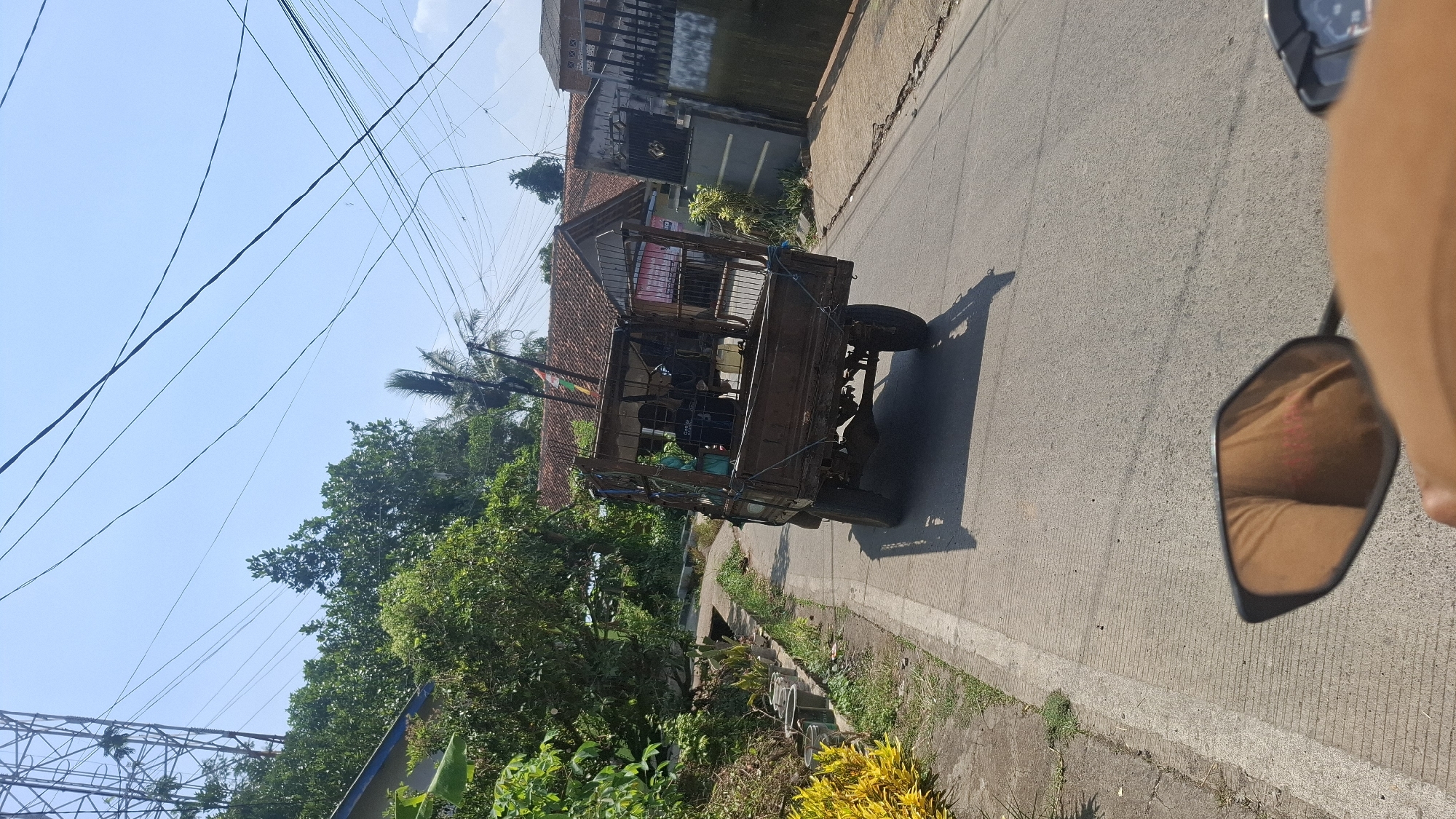 An image captured from a motorcycle, showing a weathered, three-wheeled utility vehicle driving on a paved road in Ciharashas, Indonesia. The utility vehicle, with a caged rear cargo area, carries two individuals. The driver is obscured, while a passenger wearing a black t-shirt is visible in the cage. The vehicle is laden with items, including plastic containers. The road is bordered by a concrete sidewalk and a steep, vegetated embankment on the left, featuring potted plants. On the right, residential buildings with red-tiled roofs are visible, along with a metal gate. Overhead, a complex network of power lines crisscrosses the clear blue sky, and a tall communication tower is present in the background. The perspective of the image is from behind the motorcycle's handlebars, with a portion of the rider's arm and clothing visible in the lower foreground, reflected in the motorcycle's mirror. The scene is set under bright daylight.