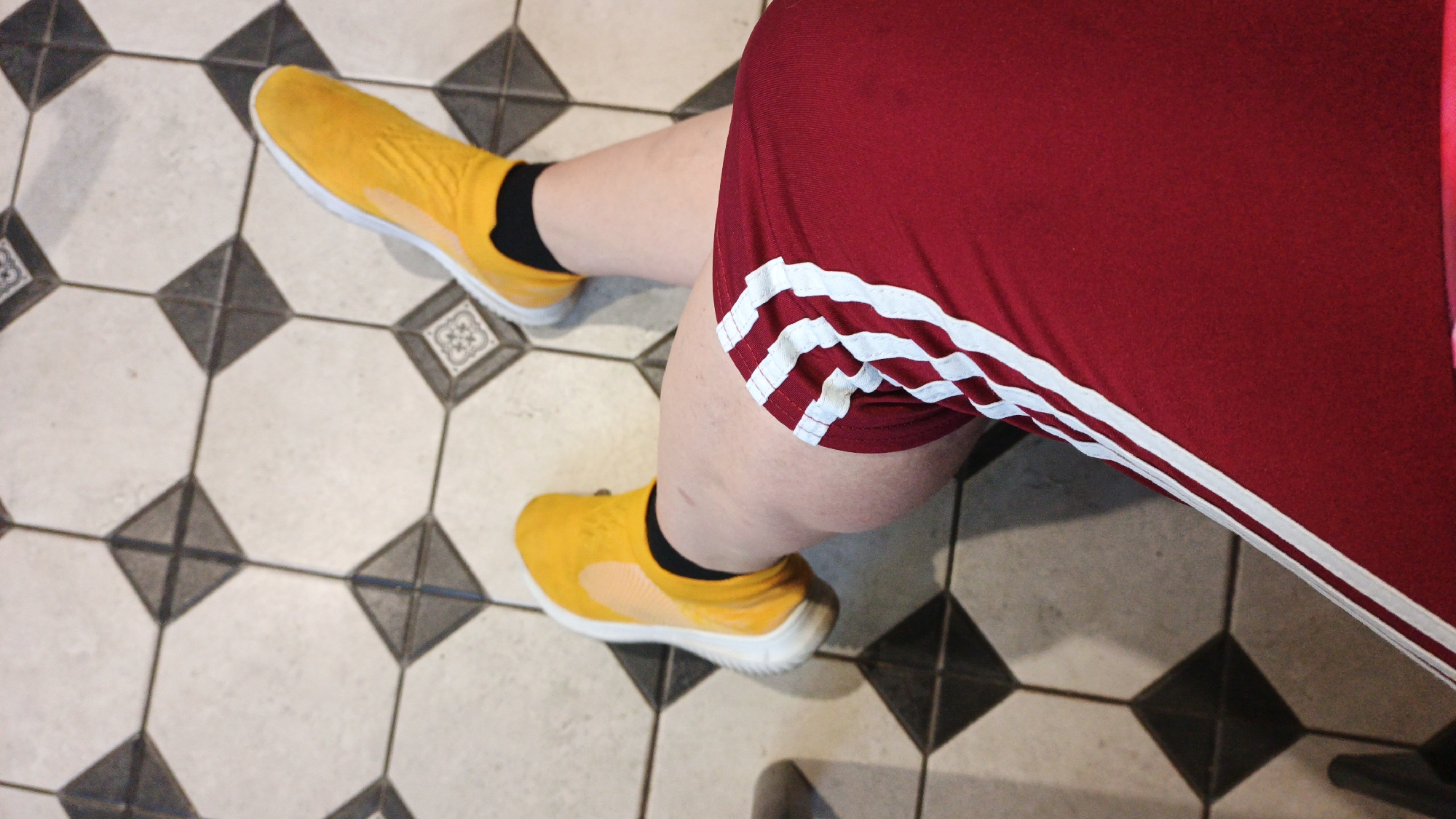 A person's legs, wearing yellow slip-on sneakers with white soles and black socks, and a red athletic skirt with white stripes, are visible. The person is sitting or kneeling on a tiled floor with a repeating pattern of white octagons and black squares. The scene is indoors. The location is the city of Drezna, Russia.