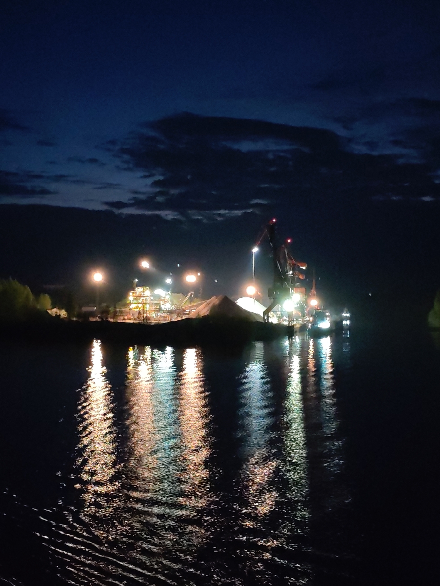 The image depicts a nighttime scene in Старое Петровское, Russia. In the middle ground, a landmass with industrial infrastructure, including cranes and buildings illuminated by bright lights, is present. A large pile of material is also visible on the landmass. The foreground features water, reflecting the lights from the infrastructure and nearby light sources, creating bright streaks. The background shows a dark sky with some clouds. No people are visible in the scene.