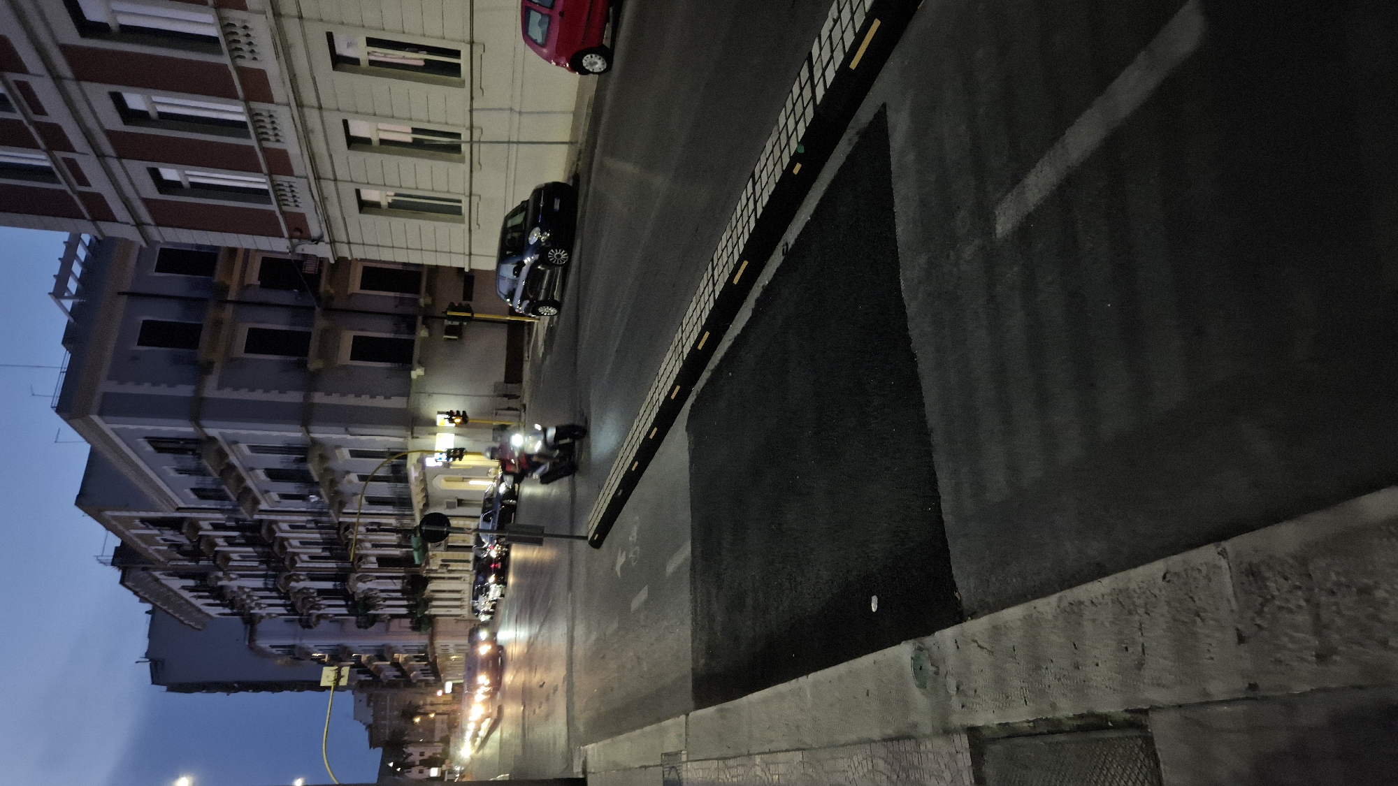 This image depicts a street scene in Taranto, Italy, at dusk. The viewpoint is from a sidewalk looking across a road. On the left, a textured concrete sidewalk and curb are visible. The road itself is paved and shows lane markings. In the foreground, a raised speed bump is installed across the road, with alternating light and dark textured sections. A small, dark patch of newly laid asphalt is situated in front of the speed bump.
Further down the road, traffic is moving. Several cars, their headlights illuminated, are traveling in both directions. A single individual is riding a motorcycle, moving away from the camera. On the right side of the road, parked cars are visible, including a dark blue hatchback and a red hatchback.
The background features multi-story buildings with classical architectural elements, characterized by multiple windows and balconies. The sky above is a deep blue, indicating twilight. Traffic lights are visible at an intersection in the middle ground. The overall impression is of an urban environment at the end of the day, with traffic and illuminated buildings contributing to the atmosphere.