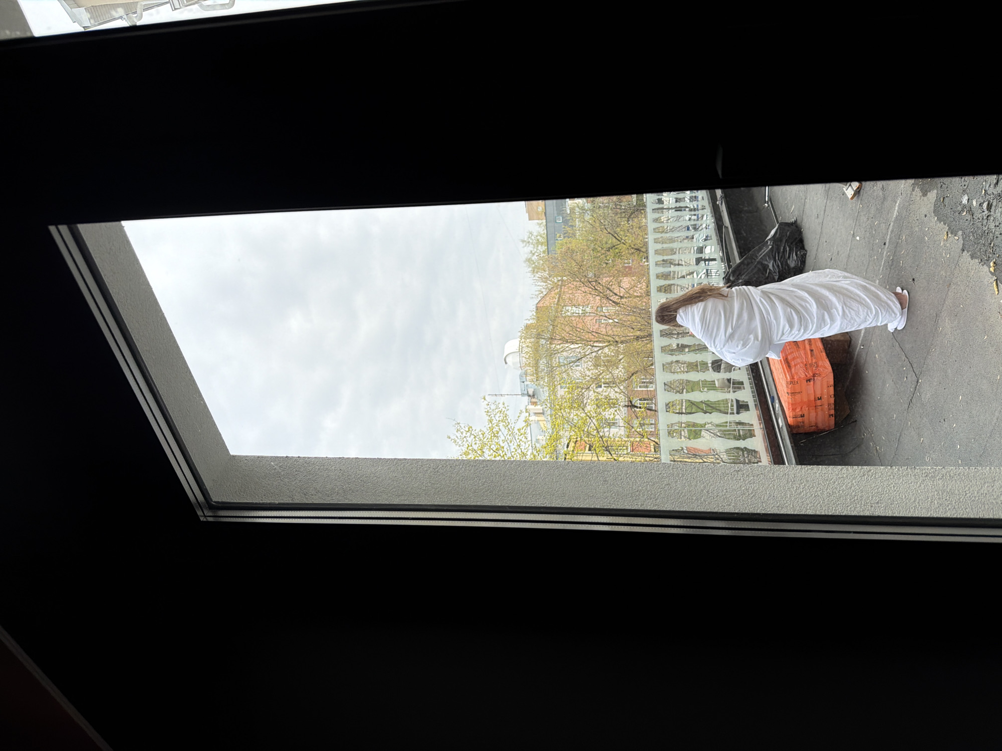 The image shows a view from inside a building, looking out through a window. Outside, a person wrapped in a white covering stands on a rooftop. The person is wearing white footwear. There are building materials next to the person, and a black plastic bag is nearby. In the background, buildings are visible, with trees showing signs of early spring foliage. The environment appears to be urban. It is impossible to specify the location within Moscow from the provided picture.