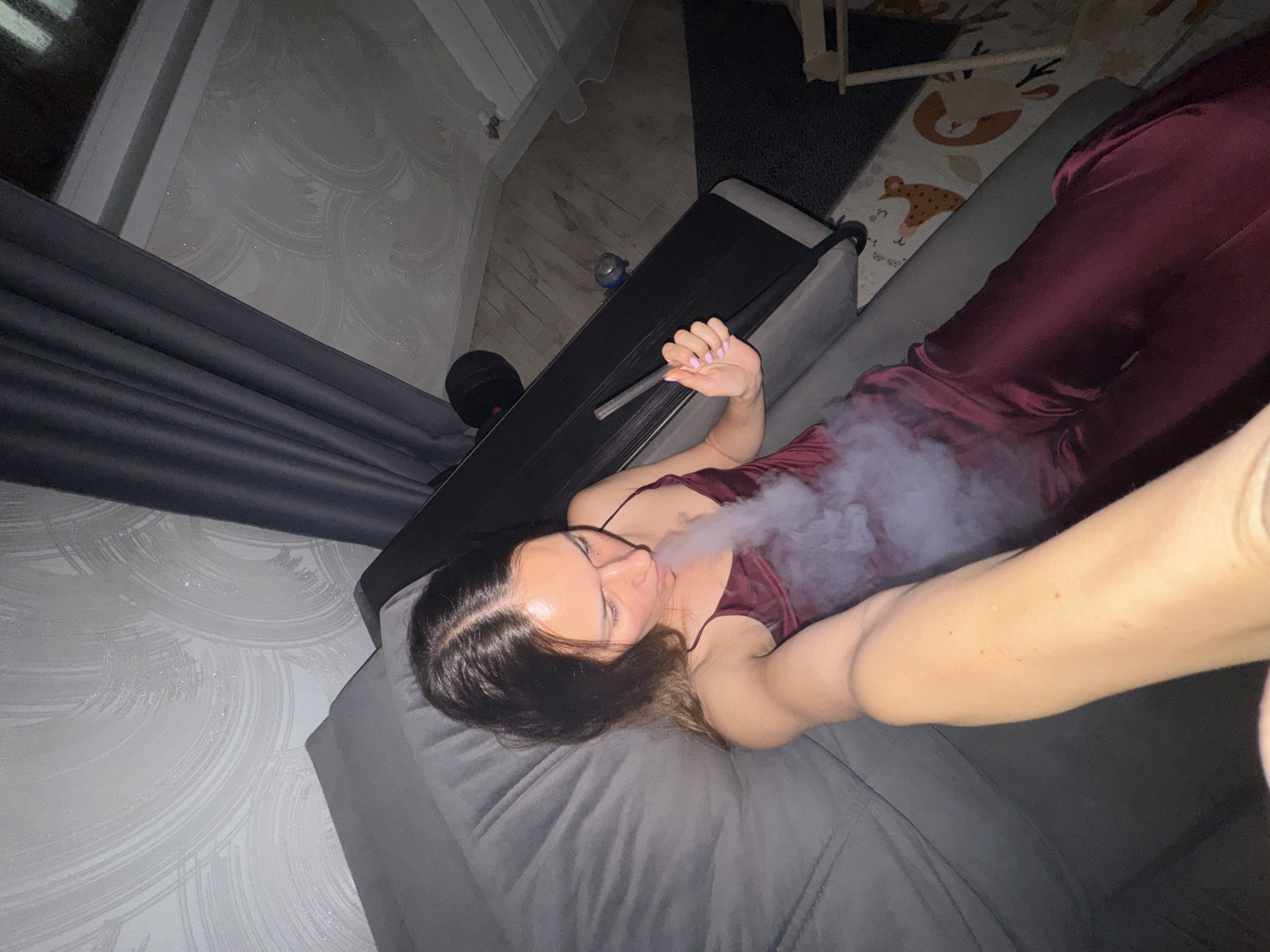 A high-angle indoor photograph depicts a young adult female from the chest up, positioned on a grey upholstered surface, likely a couch or bed. She has dark brown hair with a center part and wears a dark red satin-like garment with thin straps. The woman is exhaling a dense plume of white vapor from her mouth, holding a clear, slender pipe or mouthpiece in her right hand, which has light pink nail polish. Her arm extends towards the lower left of the frame, indicating a selfie. In the background, to the left, is a wall with light-colored wallpaper featuring a circular, textured pattern. Dark curtains are visible on the right. Further right, a dark treadmill or exercise machine is present, with a blue object resting on it. The floor beneath the treadmill has a dark patterned mat, and a lighter-colored, animal-themed patterned mat is visible to the right of the woman, with a portion of a light wooden structure also present. The scene is illuminated by artificial light, suggesting an evening or nighttime setting in Sochi, Russia.