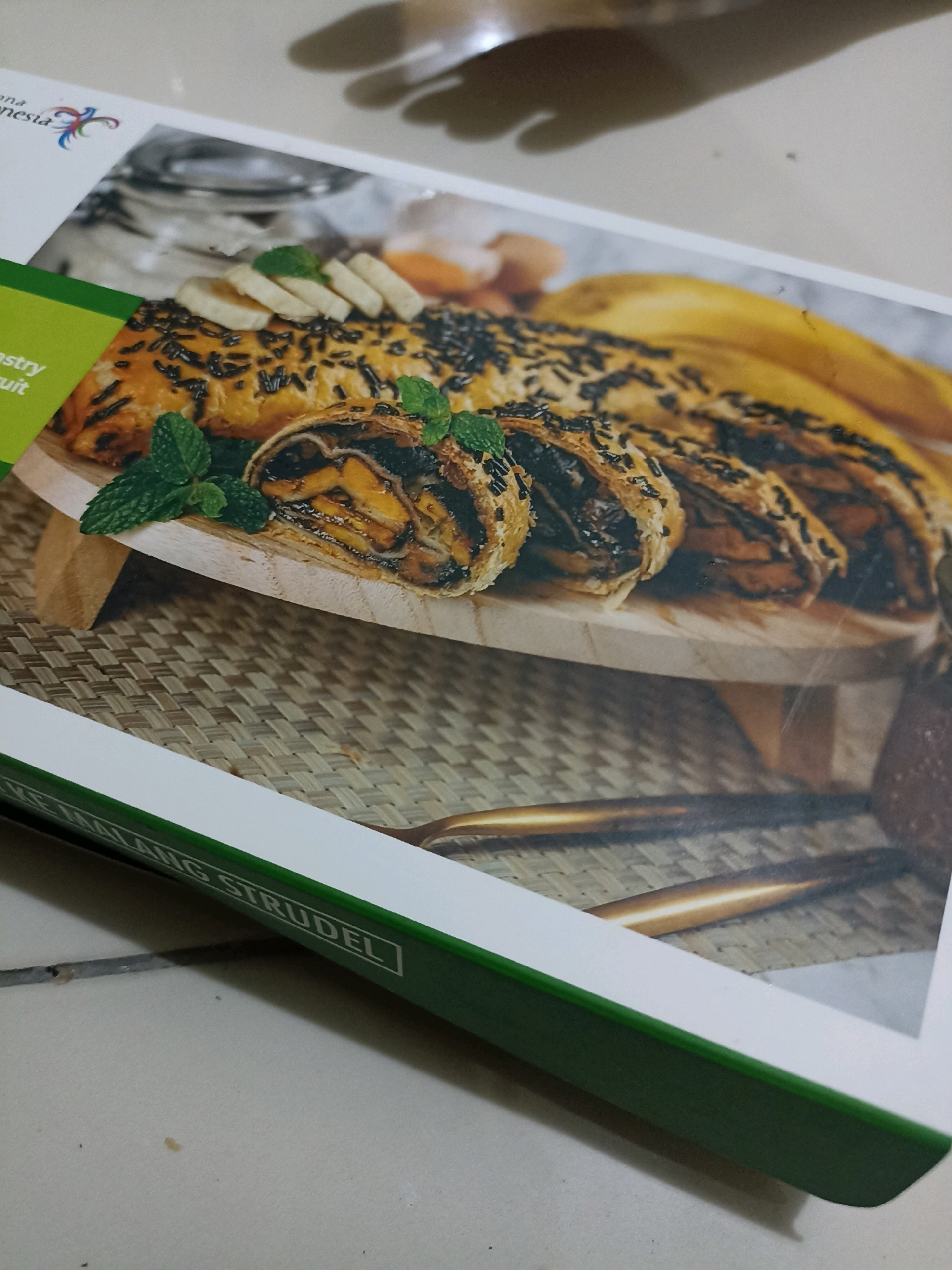 A close-up, angled shot focuses on a cookbook, with a section of its cover featuring a close-up image of a pastry. The pastry, possibly a strudel, is sliced and shows layers of dough, chocolate, and banana. It's garnished with fresh mint leaves and sprinkled with chocolate shavings. The cookbook itself has a green spine with white text that reads "KE MALANG STRUDEL", indicating it's a recipe for Malangen Strudel from Takeranklating, Indonesia. The background of the cookbook image is a woven mat and blurred bananas. The actual cookbook is resting on a tiled surface, with shadows from an unseen object cast across the upper part of the image.