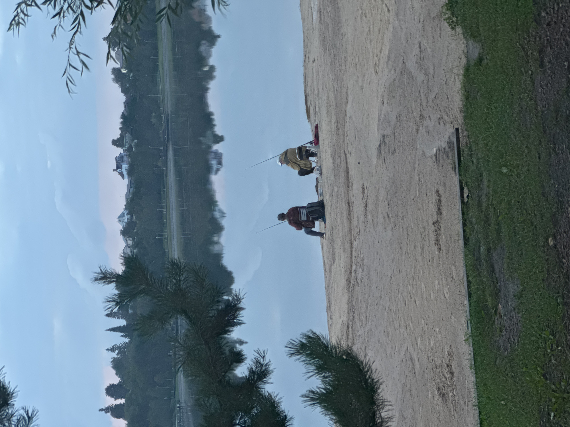 Two individuals are fishing on a sandy shore next to a calm body of water. The person on the left, wearing a maroon jacket and dark pants, is seated on a portable chair and holding a fishing rod. To their right, another individual, dressed in a tan jacket and dark pants, also sits on a chair with a fishing rod in hand. A backpack is visible behind the first fisherman, and a red object, possibly a bucket or bag, lies near the second fisherman. The environment appears to be a park or recreational area, with trees and some distant buildings lining the opposite shore, which is reflected in the still water. The sky is overcast with muted tones, suggesting an early morning or late evening setting. The foreground consists of grass and sand. The scene depicts a peaceful, leisurely activity in a natural, suburban setting.