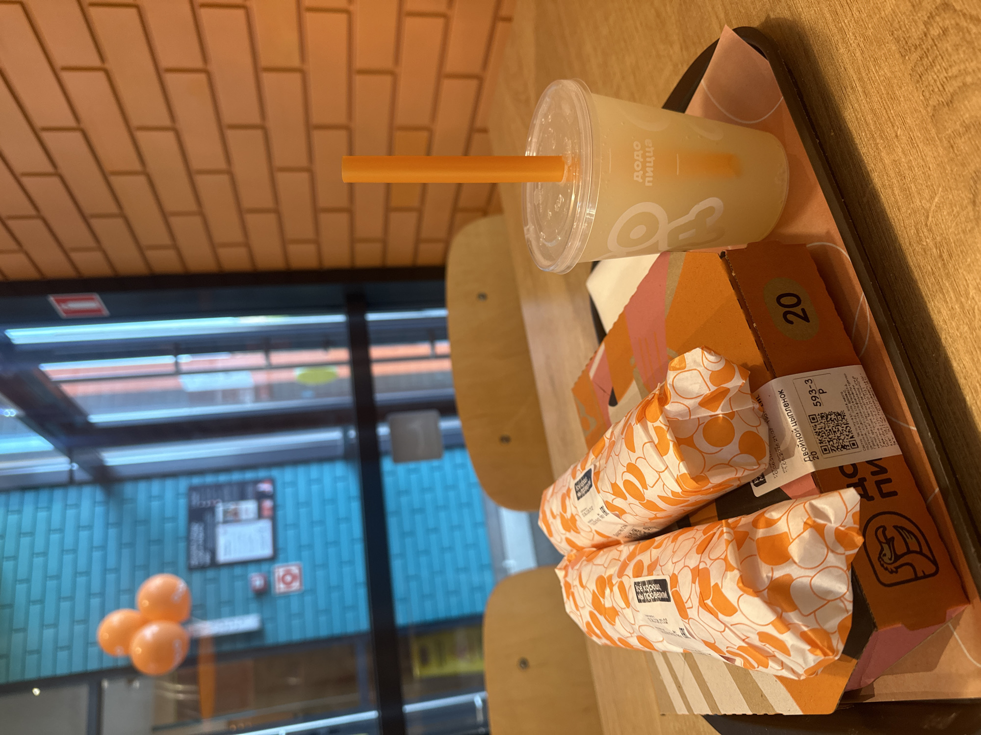 The image shows a tray with two wrapped food items, a drink in a plastic cup with a straw, and a pizza box on a wooden table. The food items are wrapped in paper with an orange and white pattern. The drink cup has a logo that appears to be "Dodo Pizza" in Cyrillic. The pizza box is orange and has a drawing of a bird on it. In the background, there is a brick wall and a large window with a view of what appears to be a street with some orange balloons. The overall scene suggests a casual dining environment, likely a fast-food restaurant. Given the "Dodo Pizza" branding, this could be a Dodo Pizza establishment. While "Dodo Pizza" has locations in Veliky Novgorod, Russia, the image itself does not contain any explicit indicators of this specific city.