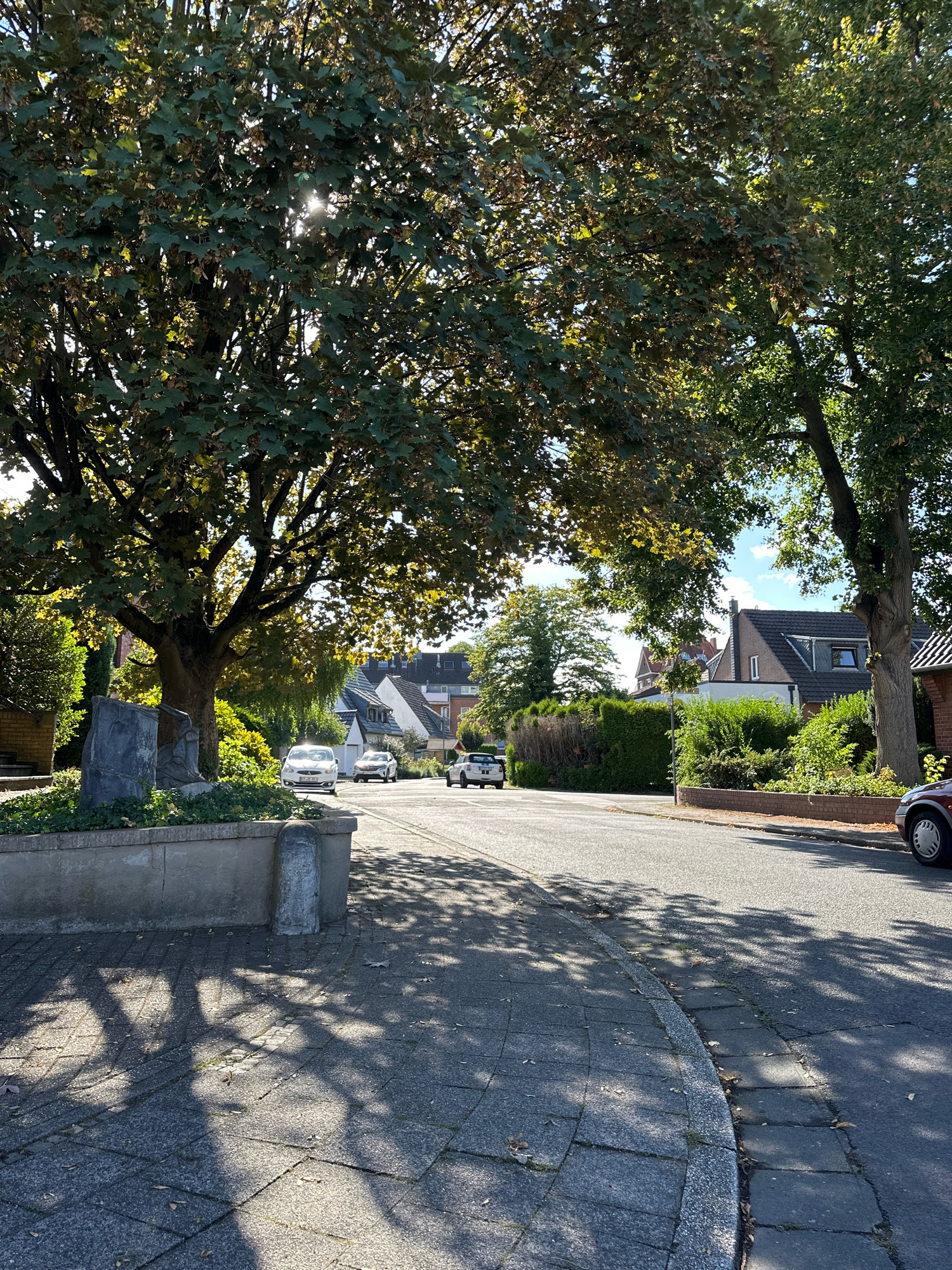 The image shows a sun-dappled street in Krefeld, Germany, lined with mature trees casting dappled shadows on the asphalt and paved sidewalk. In the foreground, a stone planter with a grey monument is visible. Several cars are parked or driving on the street, including a white sedan and a dark red vehicle partially visible on the right. Houses with tiled roofs and green hedges line the street in the background. The sky is blue with scattered white clouds.
