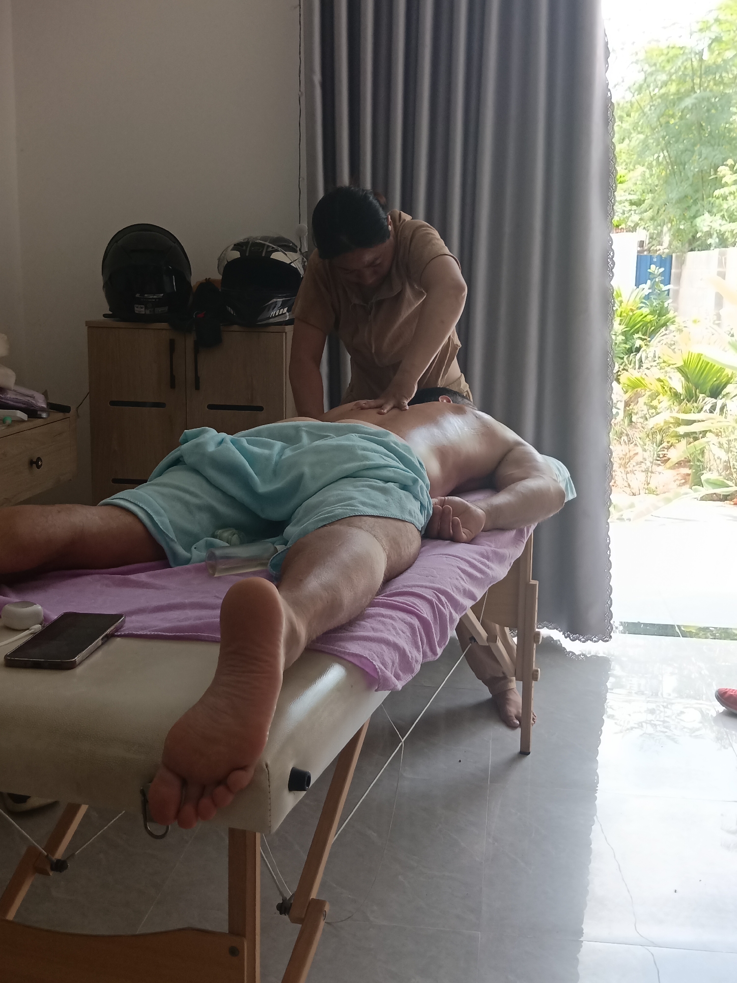 In Mũi Né, Vietnam, a person is receiving a massage on a portable massage table in a room with tiled flooring and large windows covered by grey curtains. Outside the window, lush greenery and trees are visible, suggesting a tropical or subtropical environment. The room also contains a wooden cabinet with two doors on the left, upon which two motorcycle helmets are placed. A smartphone and a white electronic device rest on the massage table near the person's feet, which are covered by a light blue towel and a pink sheet. The masseuse, wearing a tan shirt, is positioned behind the person and appears to be applying pressure to their back. The overall scene depicts a relaxing spa or therapeutic setting.