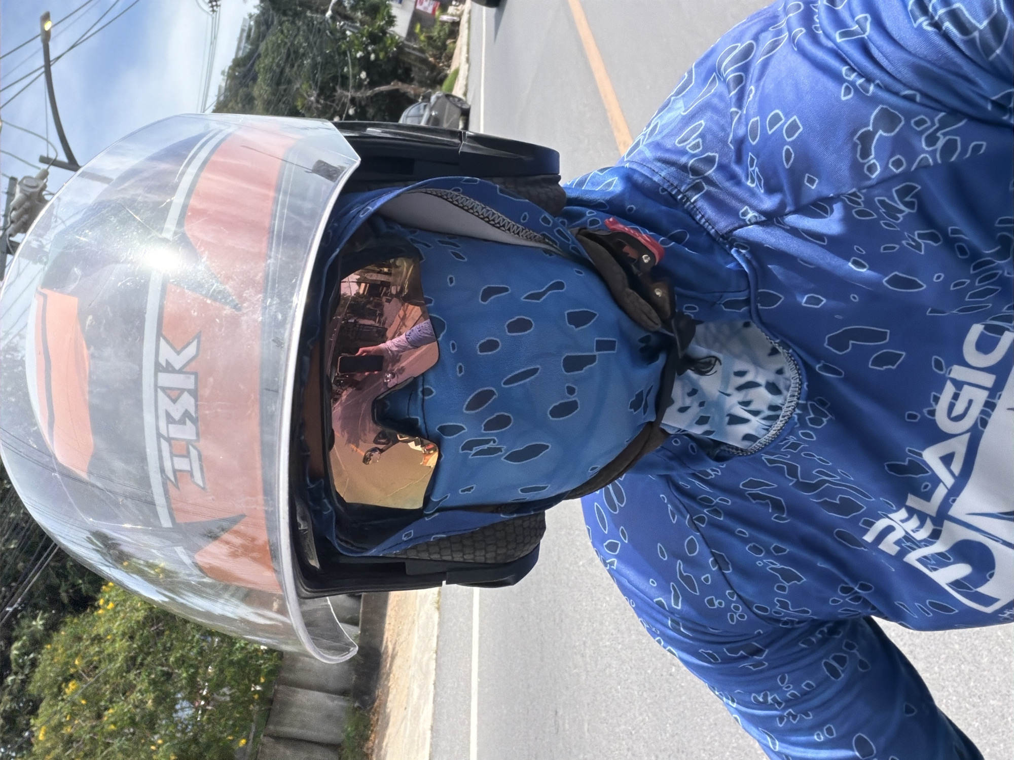 A first-person perspective shot captures a person riding a motorcycle on a paved road, likely in Baan Plai Laem, Thailand, due to the style of road markings and surrounding vegetation. The rider is wearing a full-face helmet with an orange and black design, a clear visor, tinted goggles, and a blue patterned face mask and shirt. The shirt has a logo that appears to be "PELAGIC" and a distinctive pattern of irregular shapes with dark blue outlines on a lighter blue background. The reflection in the goggles shows a person holding a phone, likely taking the selfie, and a glimpse of a street scene with buildings and trees. The road is marked with a yellow line and a white line, and there is a concrete barrier and greenery along the side. The sky is overcast.
