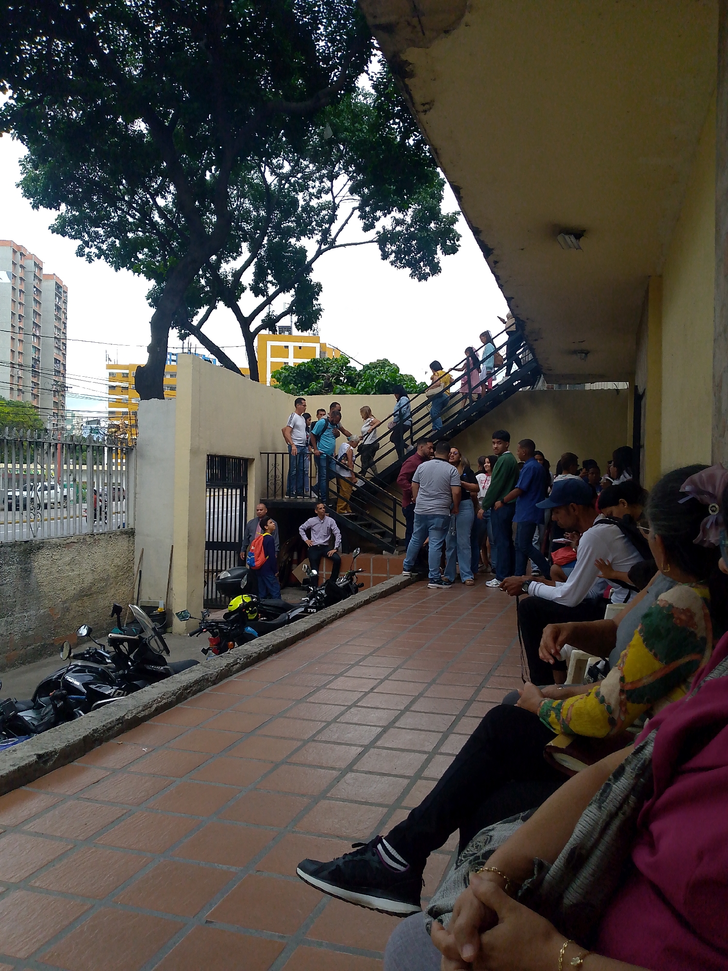 The image shows a group of people gathered on a tiled walkway in what appears to be an urban setting. A building with a yellow exterior runs along the right side of the frame, providing a covered area where several people are seated. Some individuals are standing and conversing near a staircase that leads to an upper level. The people are diverse in age and attire.
To the left of the walkway, a row of motorcycles is parked against a low wall. Beyond the wall, there is a fence and a view of buildings in the background, including high-rise apartments and other urban structures. A large tree dominates the upper-left corner of the image, partially obscuring the sky.
The scene has the appearance of a waiting area or gathering place, possibly outside a government building or service center. Based on the architectural style, weather, and context, the location is plausibly the city of Caracas, Venezuela.